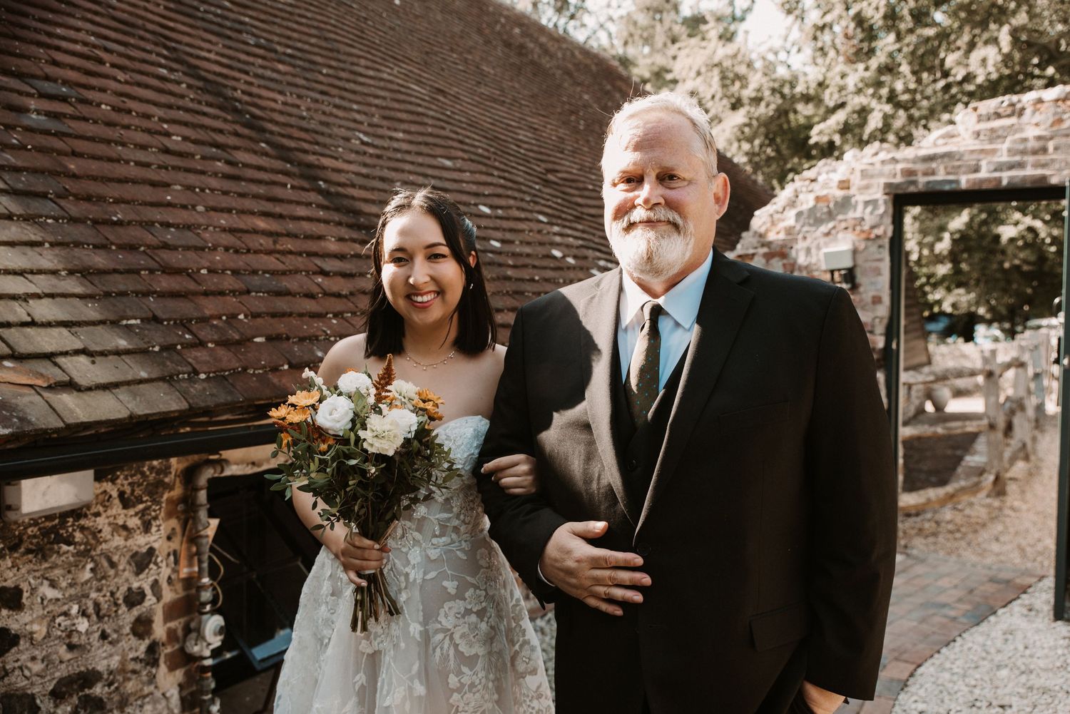 Bride in lace gown holds bouquet while walking with older gentleman in black suit at rustic venue.
