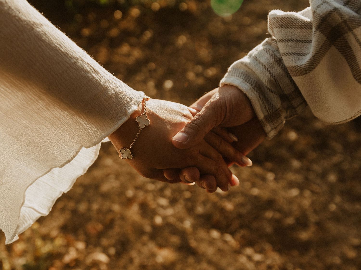 Close-up of a couple holding hands outdoors, one wearing a bracelet, surrounded by autumn leaves.