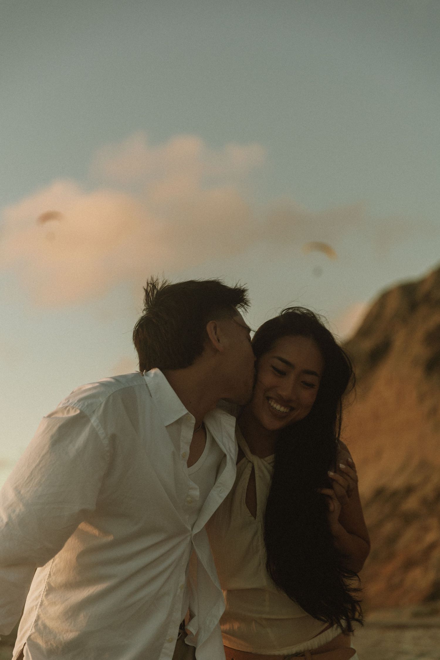 A couple shares a romantic moment at sunset, man kissing woman's cheek as she smiles joyfully on a beach.