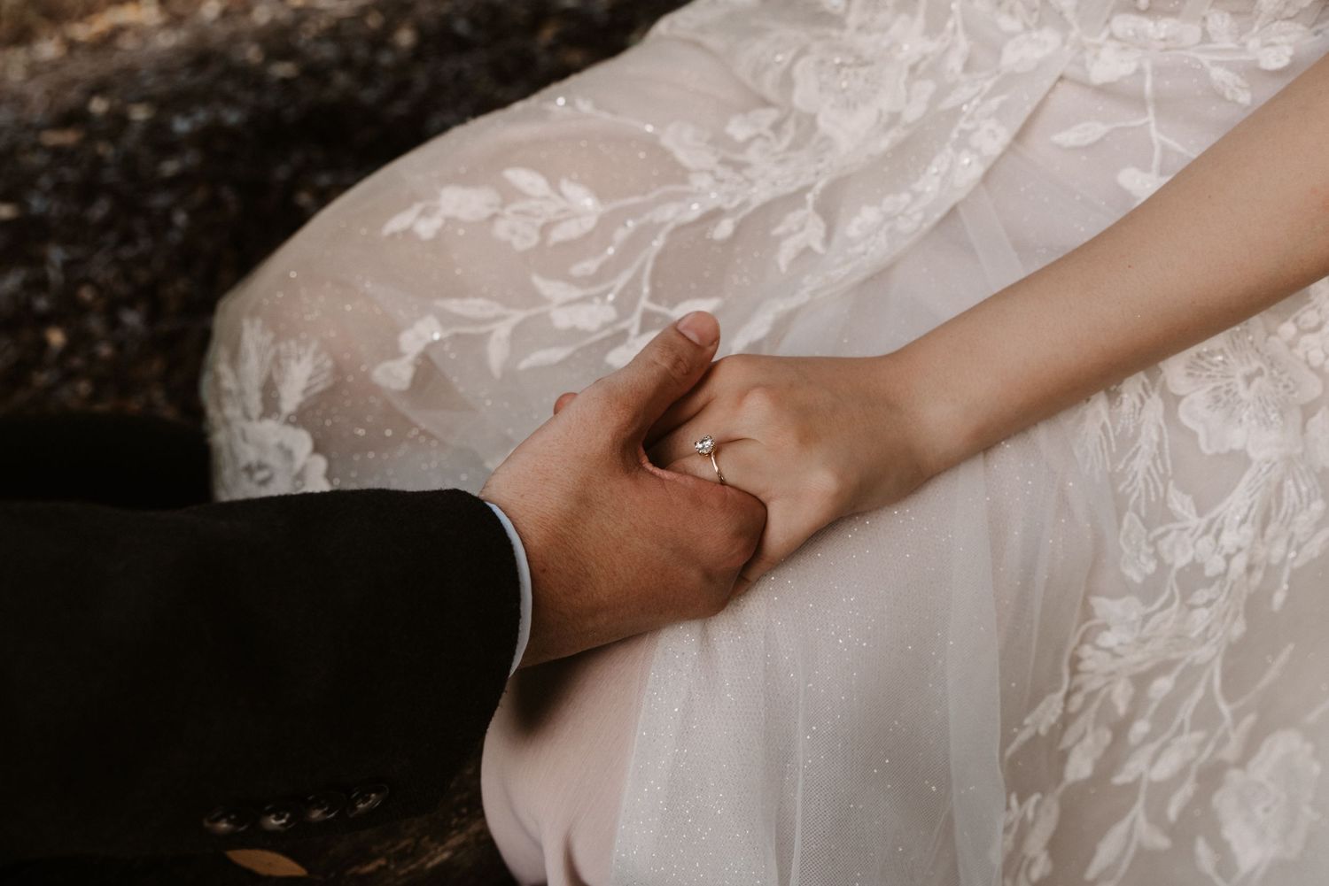 Close-up of bride and groom holding hands, wedding ring visible, bride in lace gown.