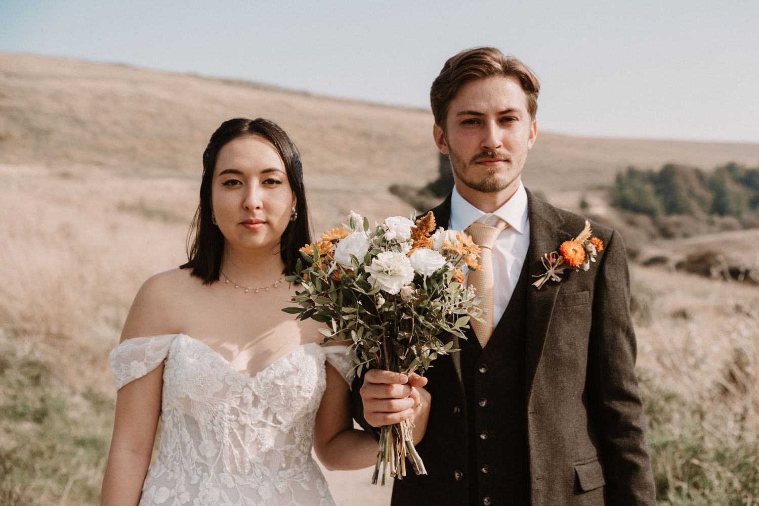Bride and groom portrait in golden field, bride holding autumn bouquet in off-shoulder lace gown.