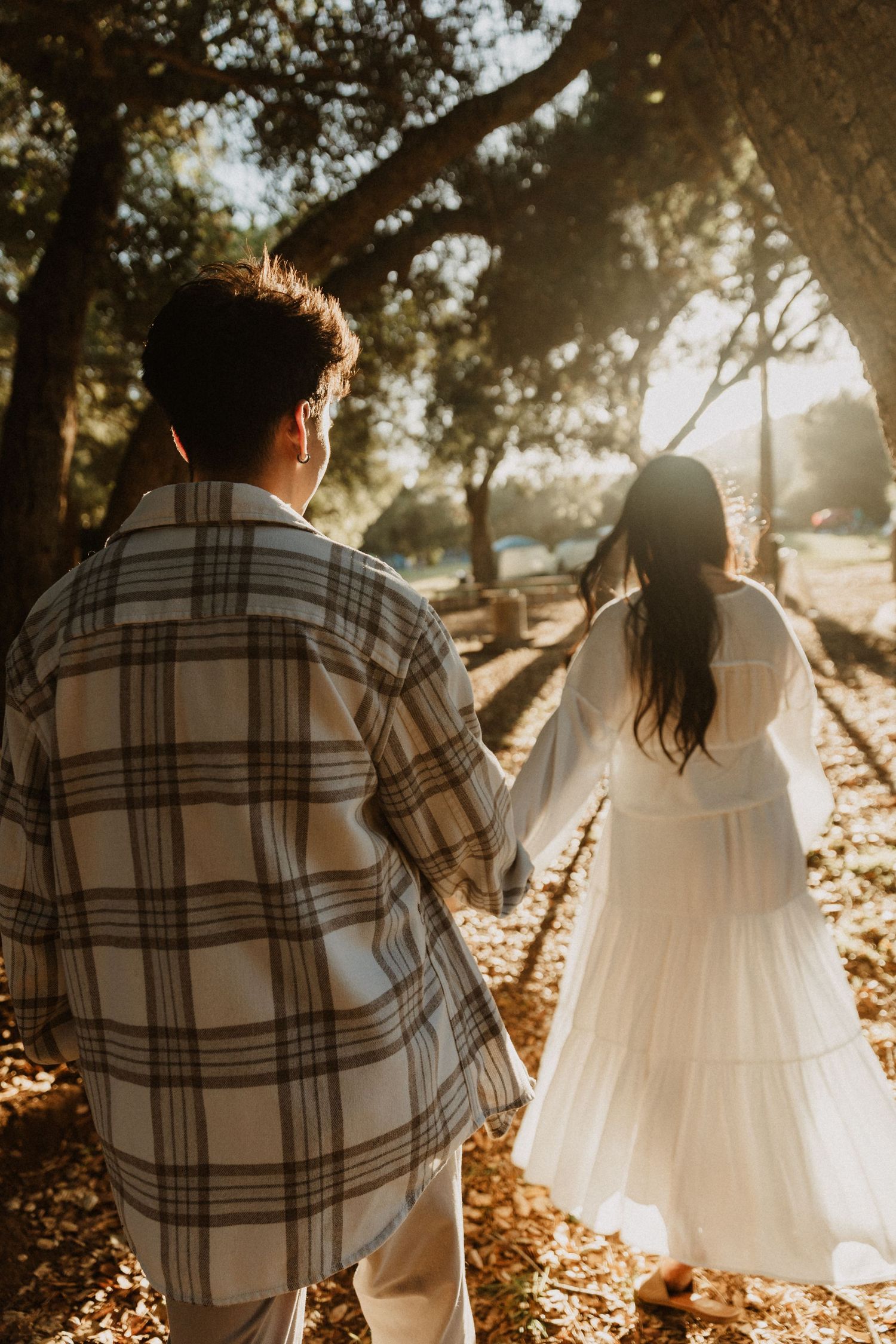 Couple holding hands walking through sunlit forest path, woman in white dress and man in plaid shirt.