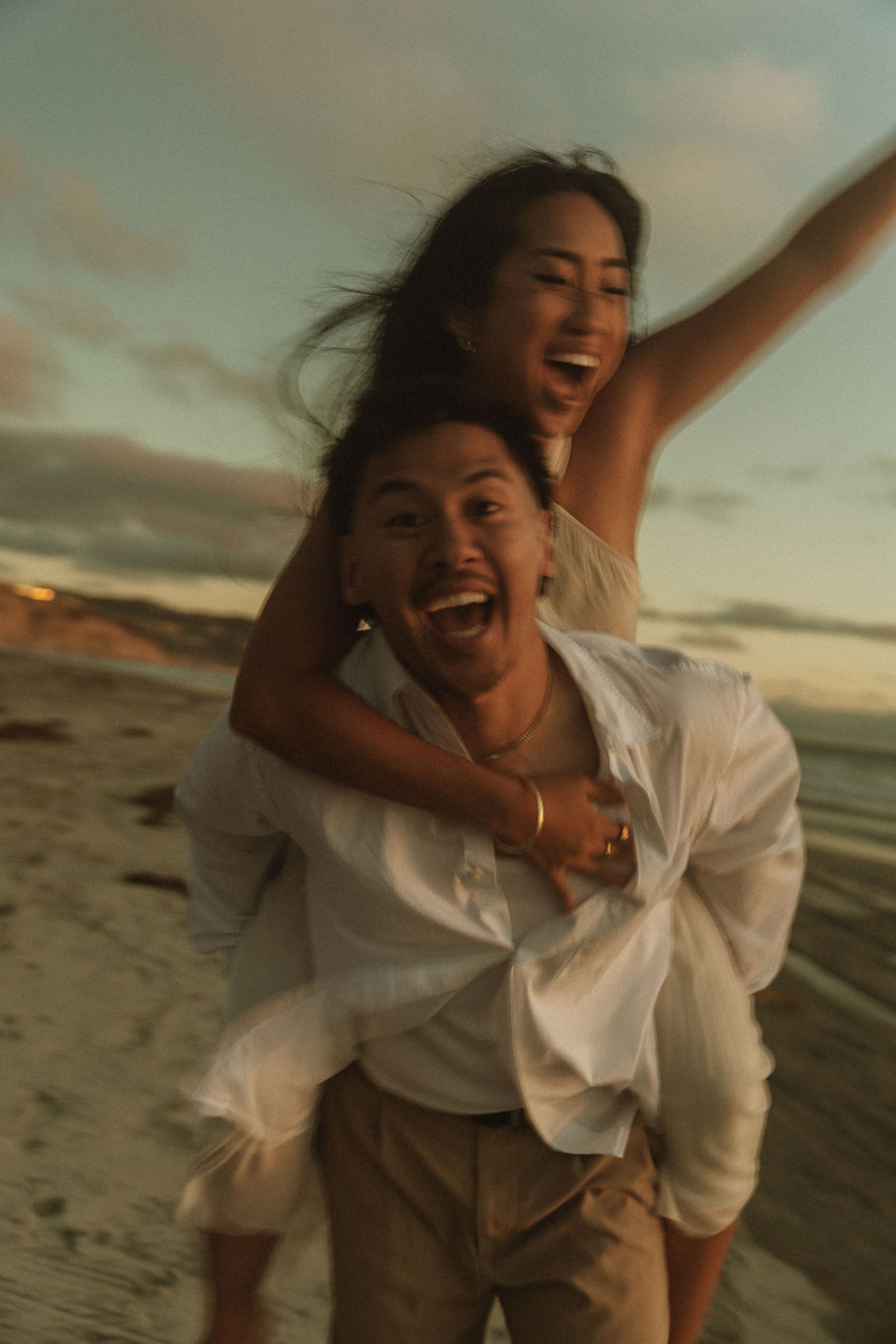 Joyful couple laughing on beach at sunset, woman on mans back with arms raised, both wearing white outfits.