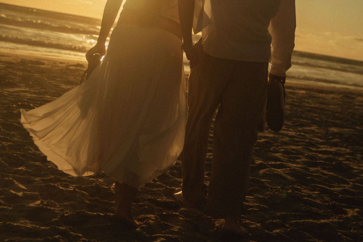 Couple walking hand in hand on a beach at golden sunset, woman in flowing white dress, barefoot on sand.