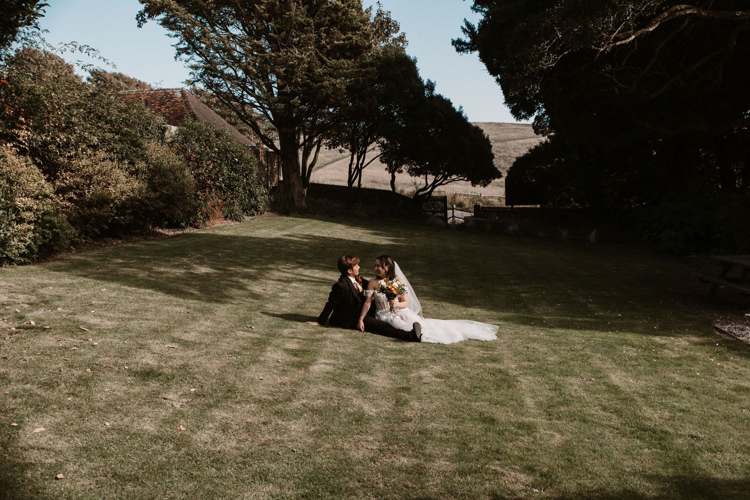 Bride and groom sitting together on green lawn surrounded by trees on sunny wedding day.