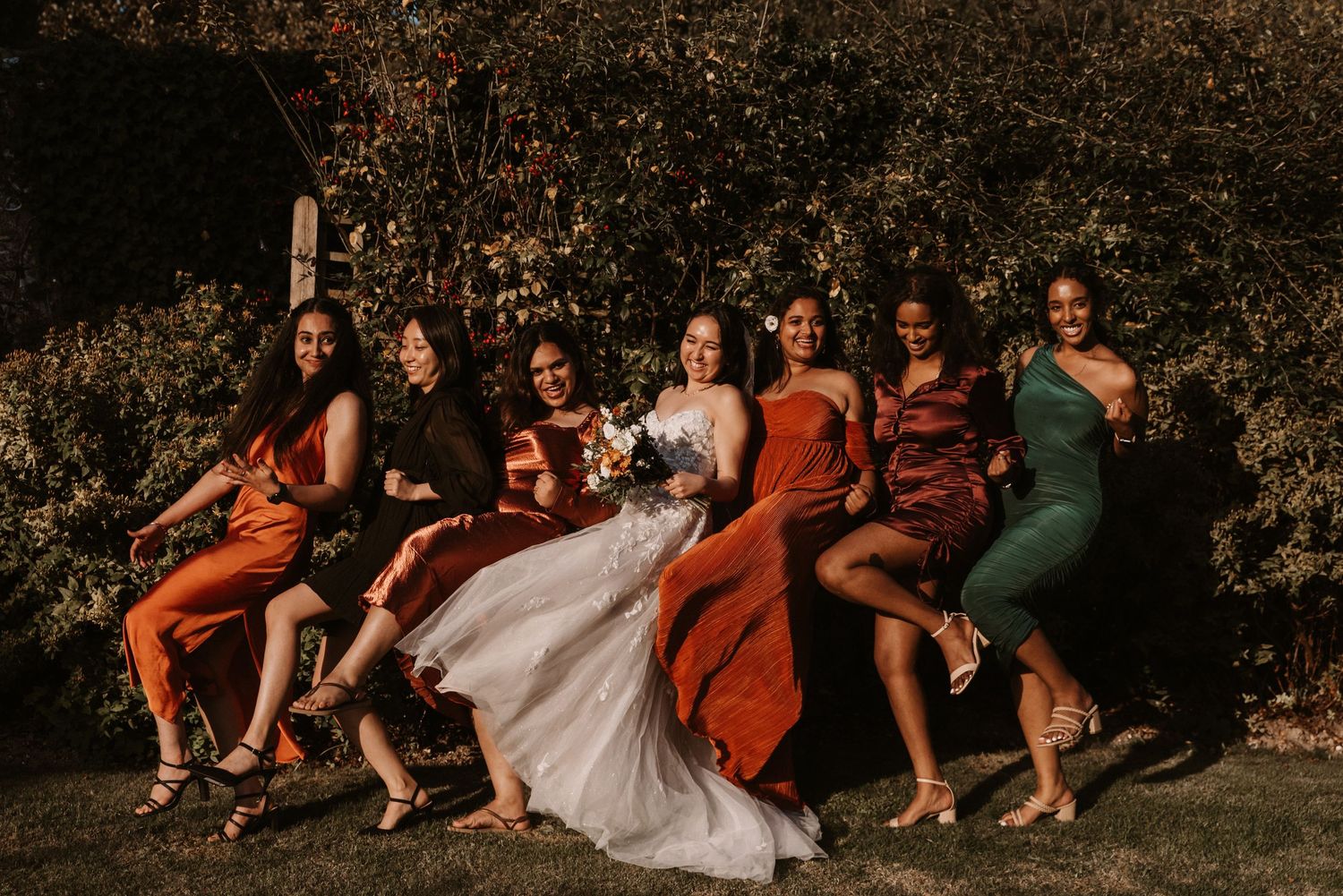 Bride surrounded by six bridesmaids in colorful dresses posing outdoors.