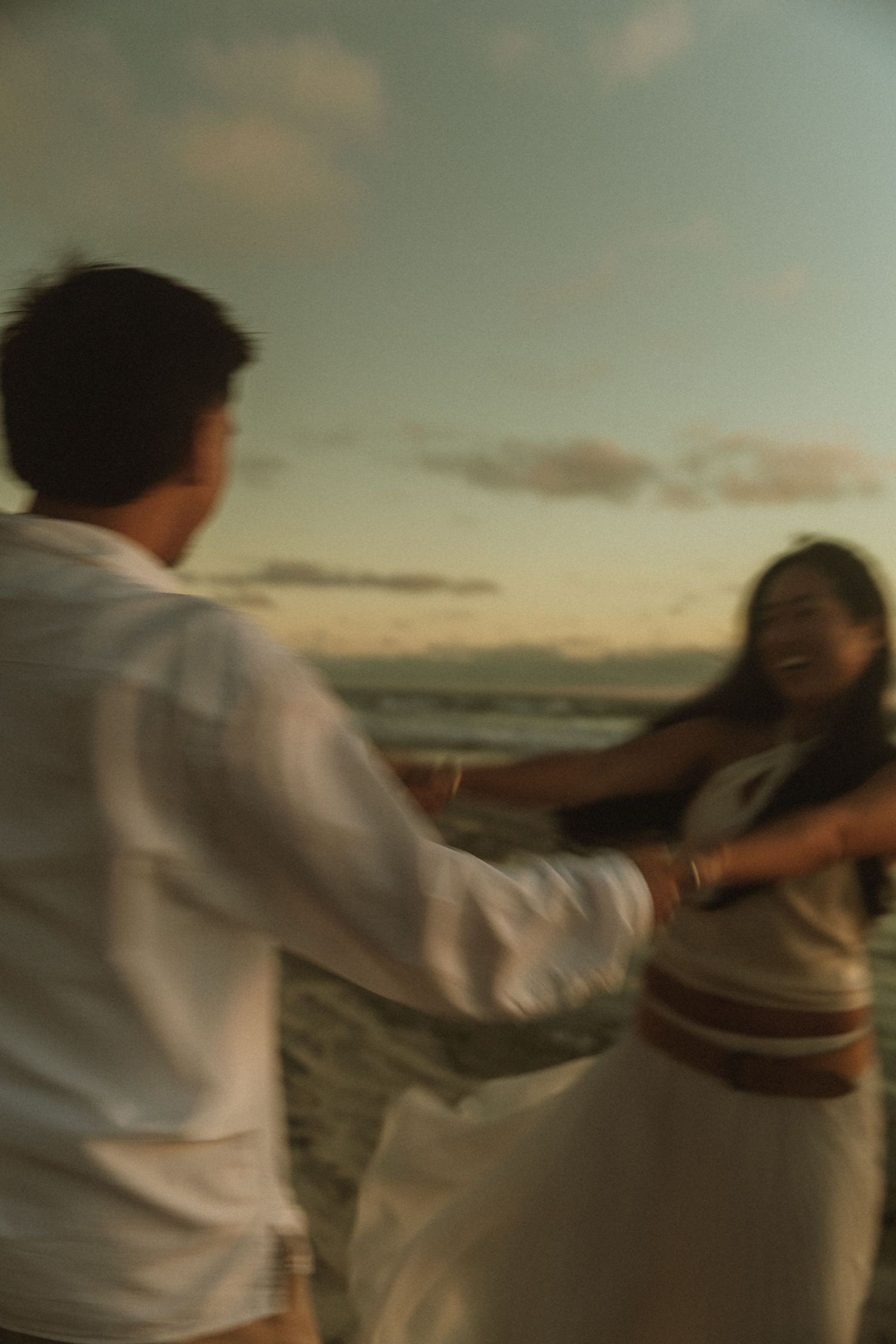 A couple dances joyfully on a beach at sunset, both dressed in white, with ocean waves in the background.