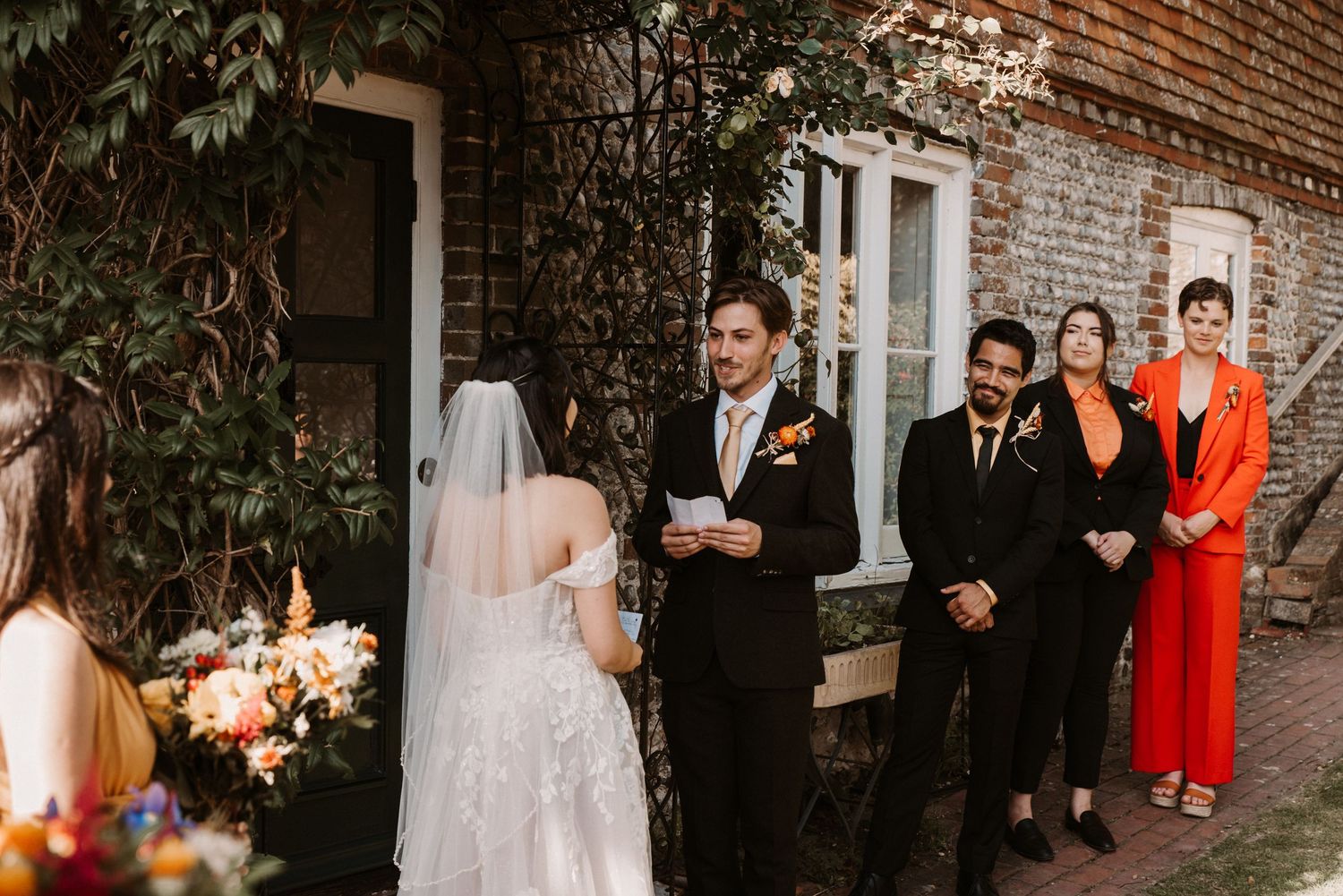 Wedding ceremony at brick venue with bride, groom reading vows, and guests in colorful attire standing nearby.