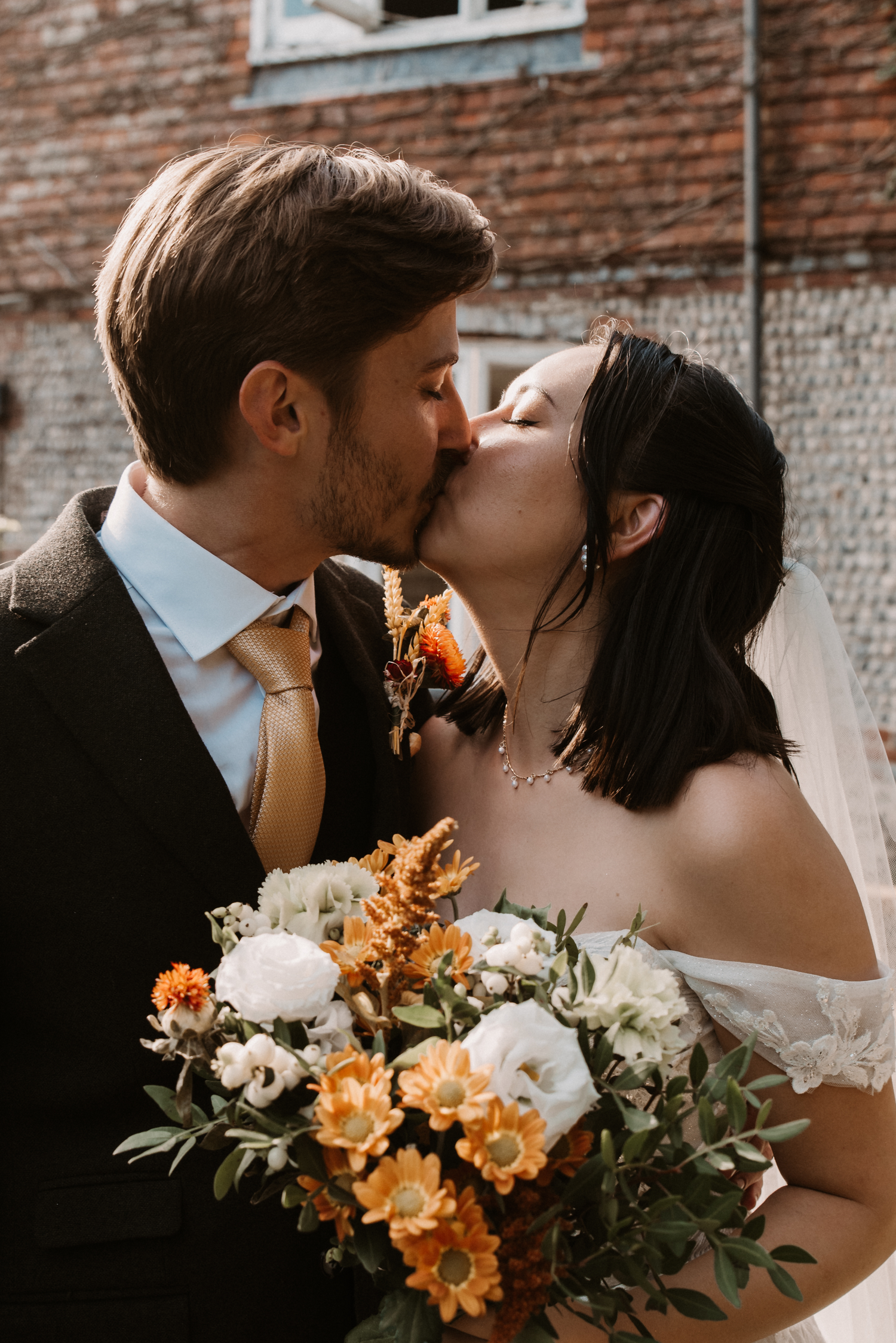 A bride and groom share a romantic kiss outdoors, with the bride holding a beautiful bouquet of orange and white flowers.