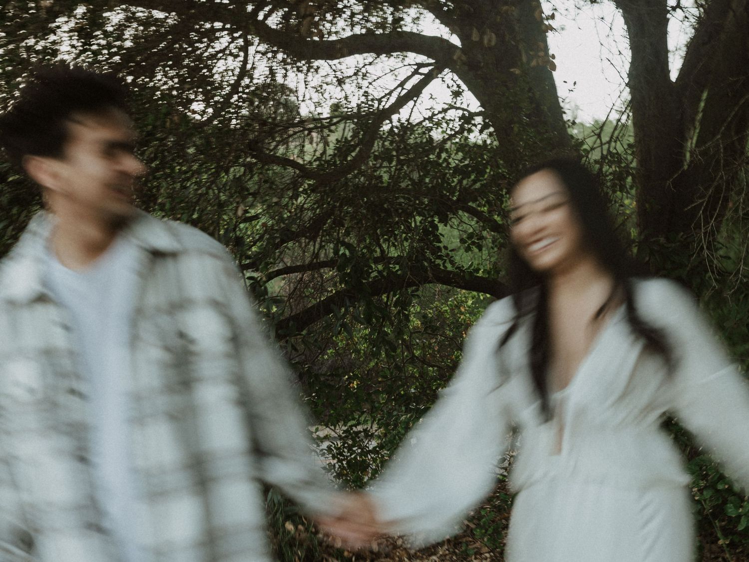 Blurry couple holding hands in a lush green forest, woman in white dress smiling, man in plaid shirt.