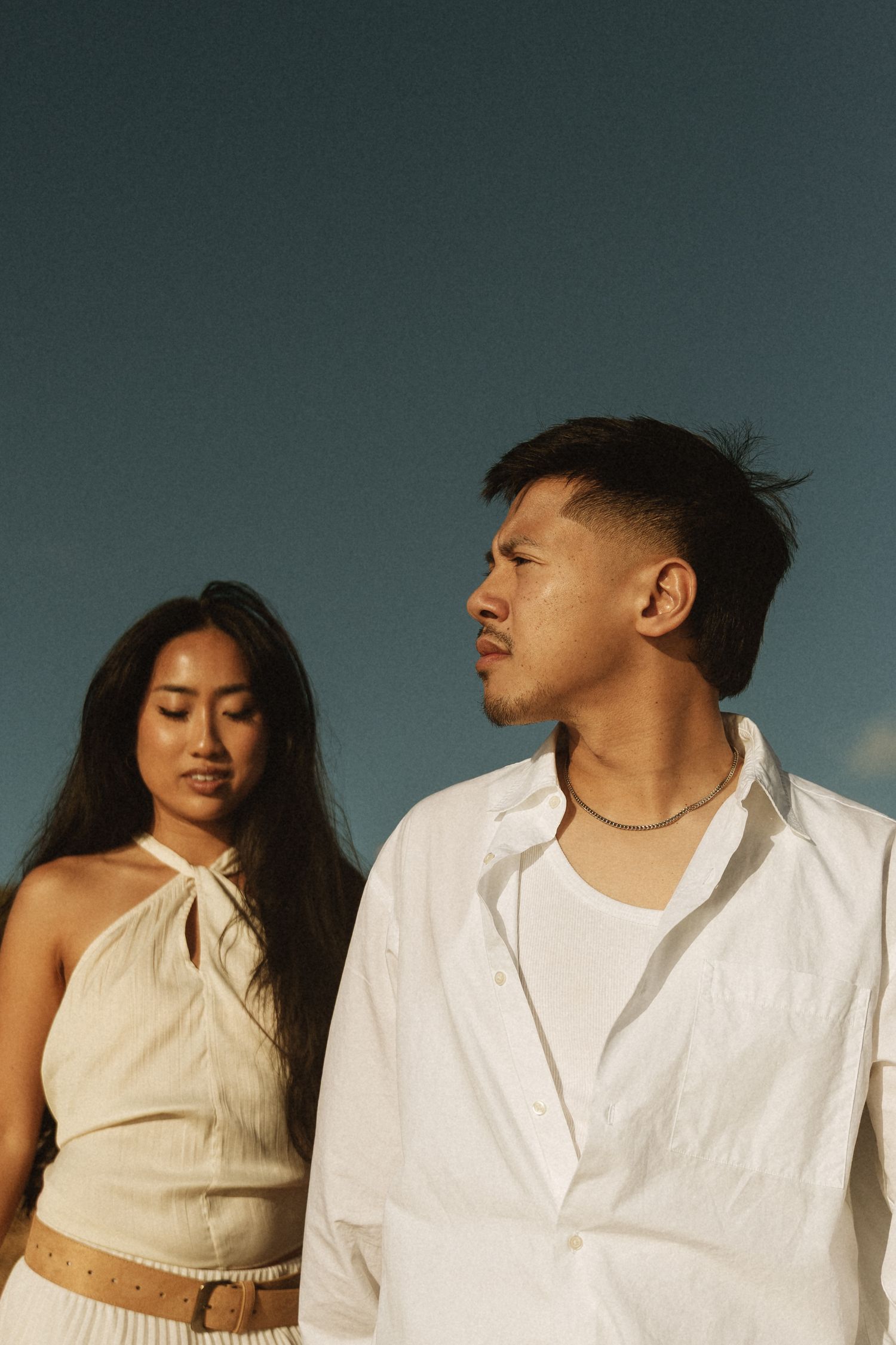 A young Asian couple in white outfits pose against a clear blue sky in warm golden light.