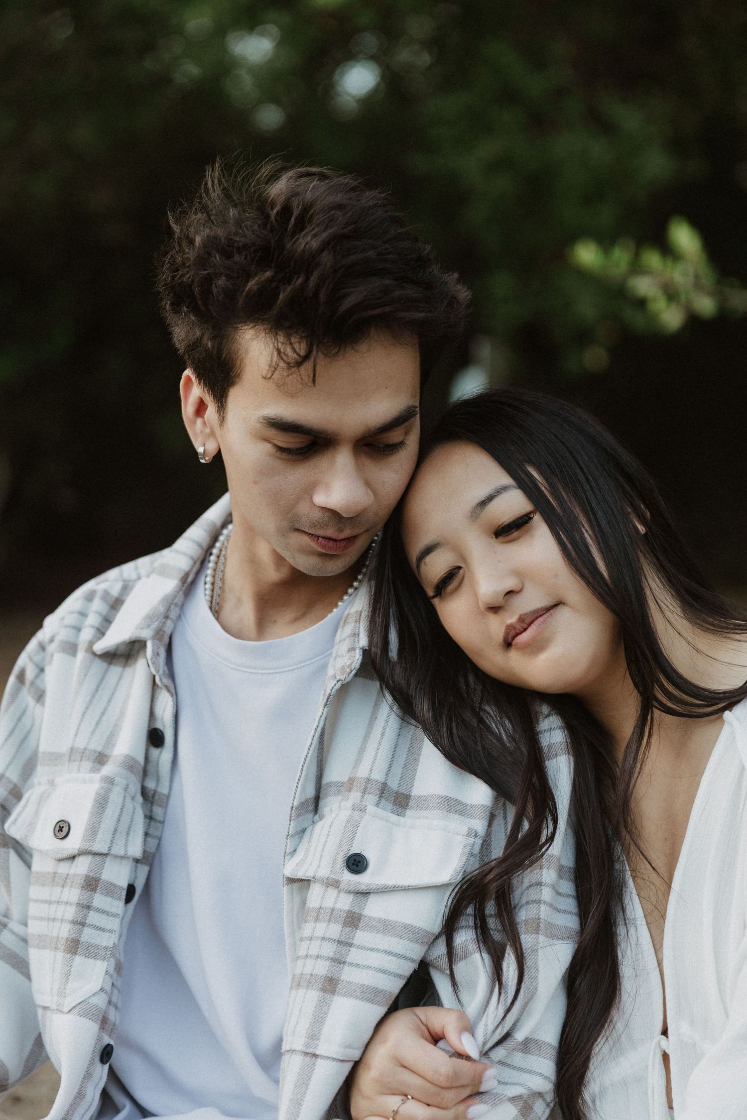 A young couple embracing outdoors, woman resting head on man's shoulder, both wearing casual light-colored clothing.