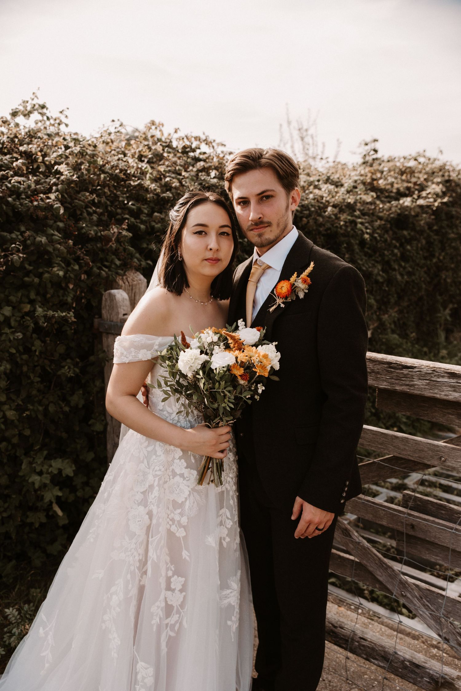 Bride and groom posing together holding floral bouquet in front of lush green garden hedge.