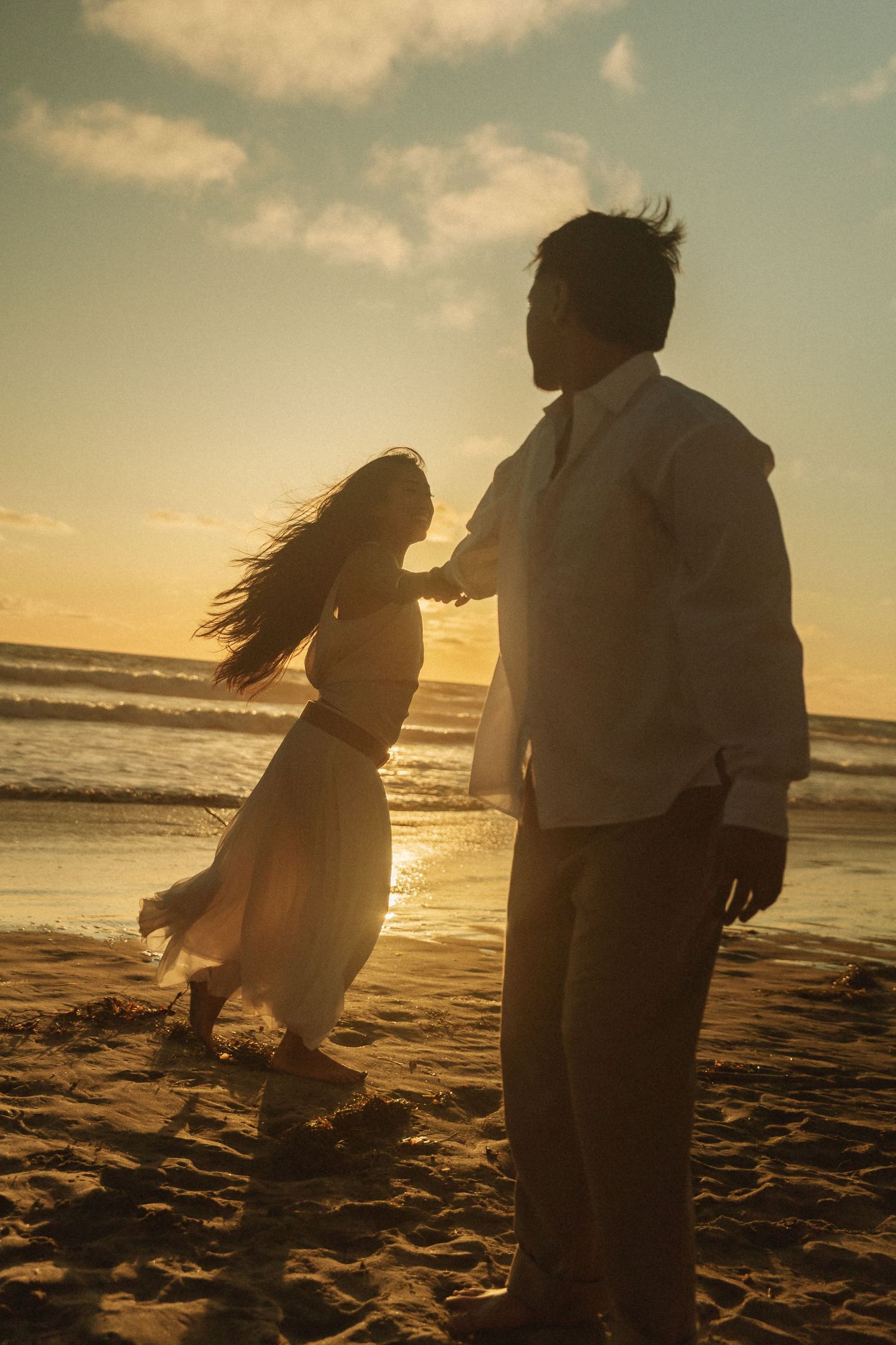 Couple silhouetted against a golden sunset on a sandy beach, woman in flowing dress with wind-blown hair.
