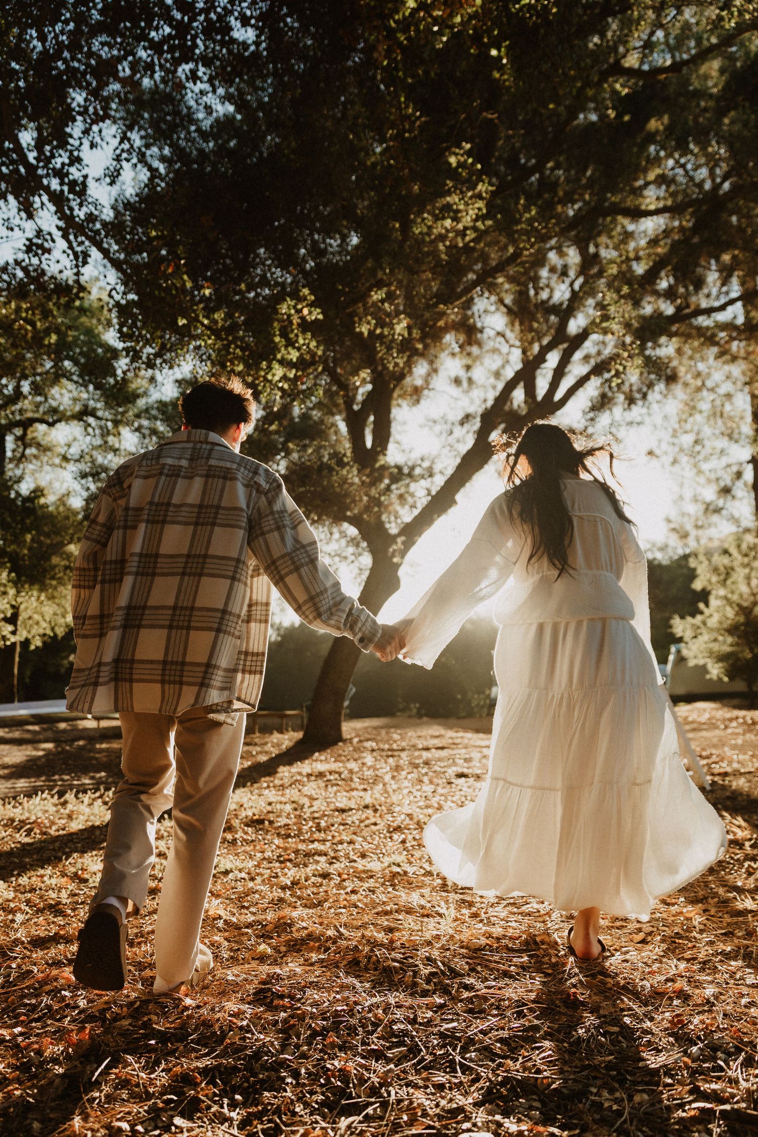 Couple holding hands walking through a sunlit autumn park, woman in white dress and man in plaid shirt.