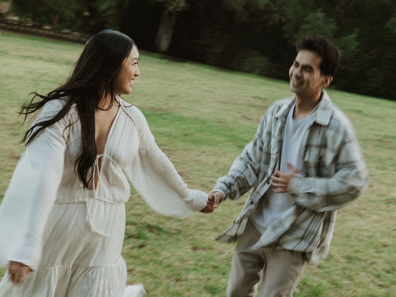 A couple holds hands and laughs while running through a lush green park, woman in white backless dress.