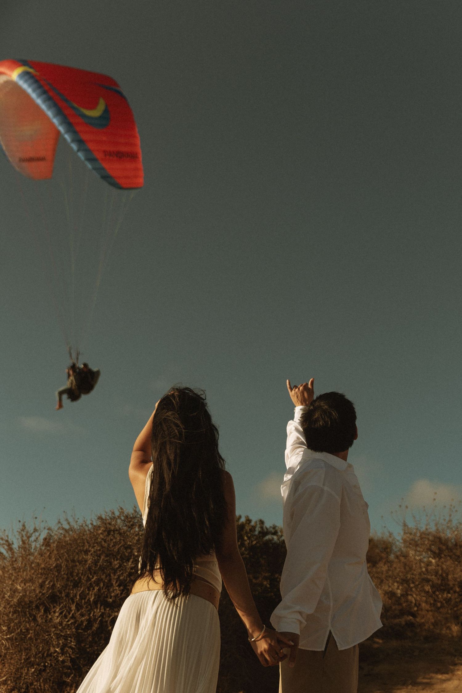 Couple in white attire holding hands, pointing at a colorful paraglider soaring in a clear blue sky above scenic shrubs.