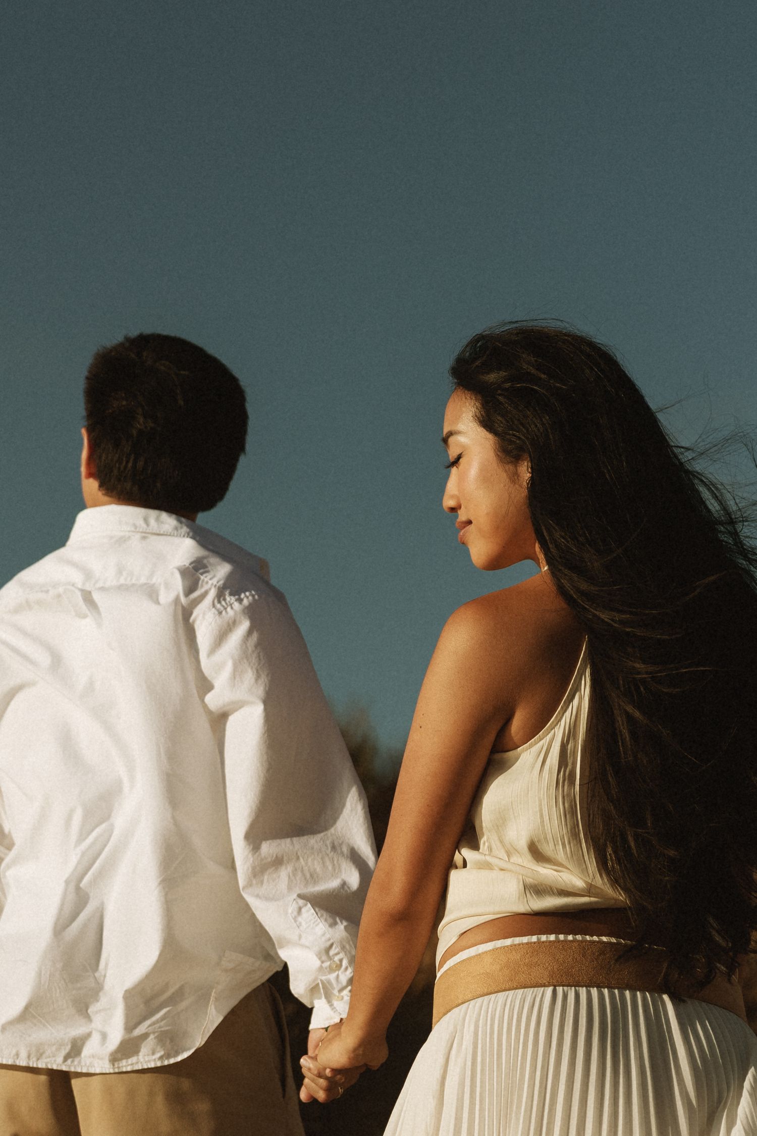 A couple holds hands against a clear blue sky, the woman in a white pleated skirt with long flowing hair.