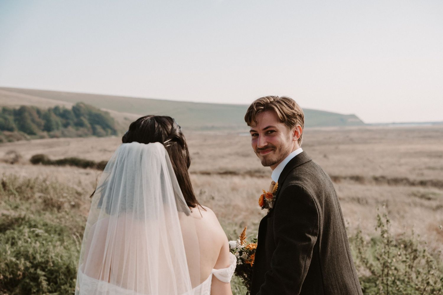 Smiling groom glances back at camera beside bride in veil with coastal field background.