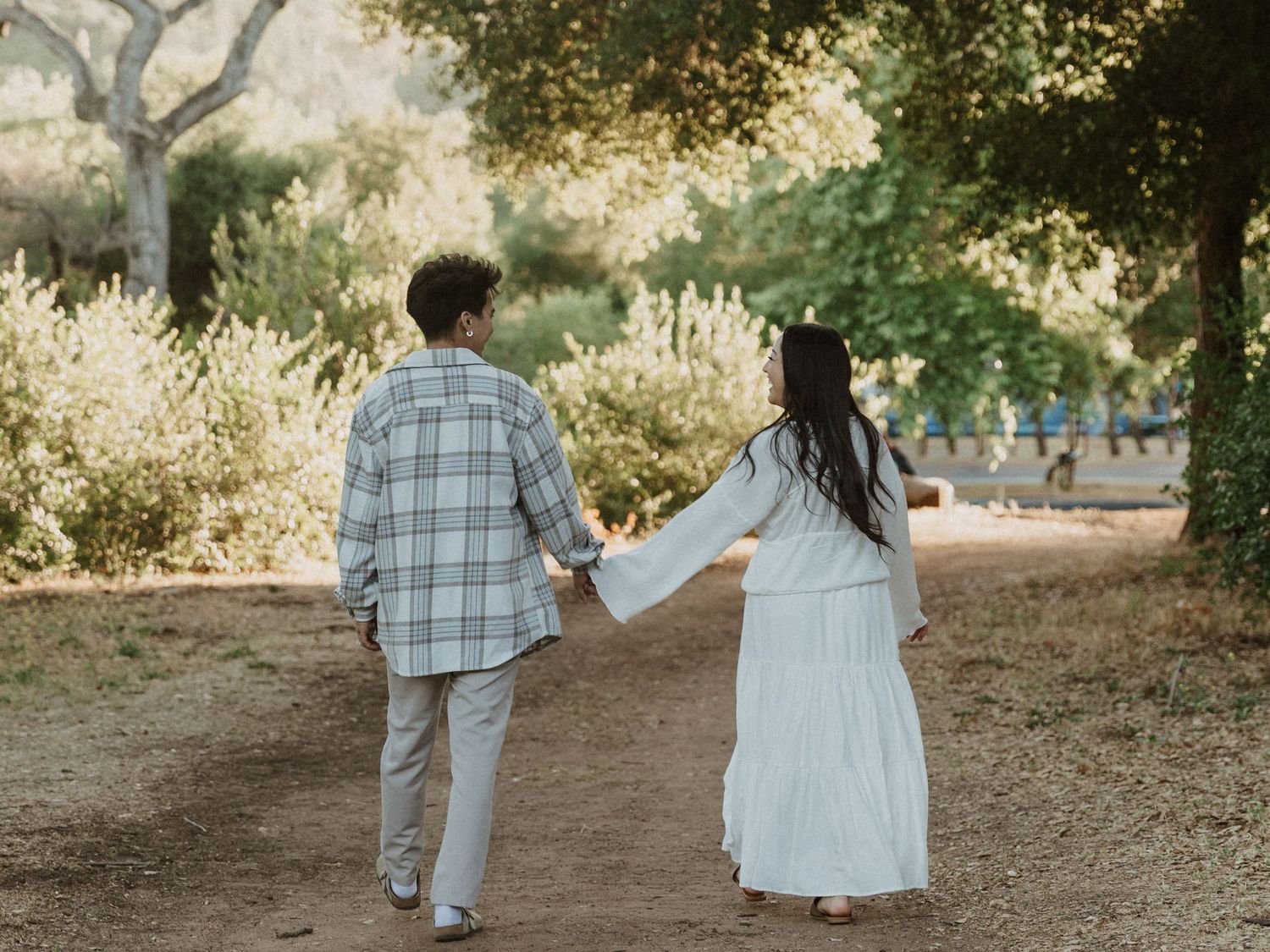 Couple holding hands walking on a dirt path surrounded by lush green trees in a sunny outdoor setting.