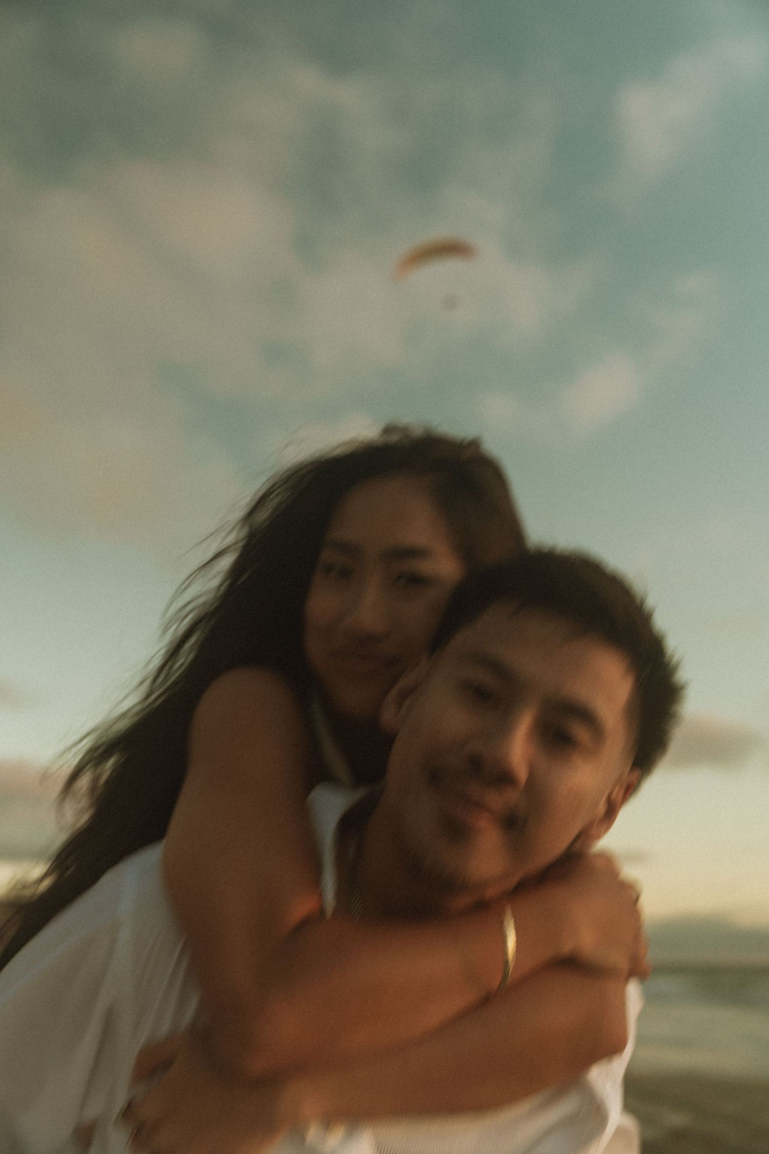 A couple shares a tender piggyback embrace at sunset, with the woman wrapping her arms around the man's neck outdoors.