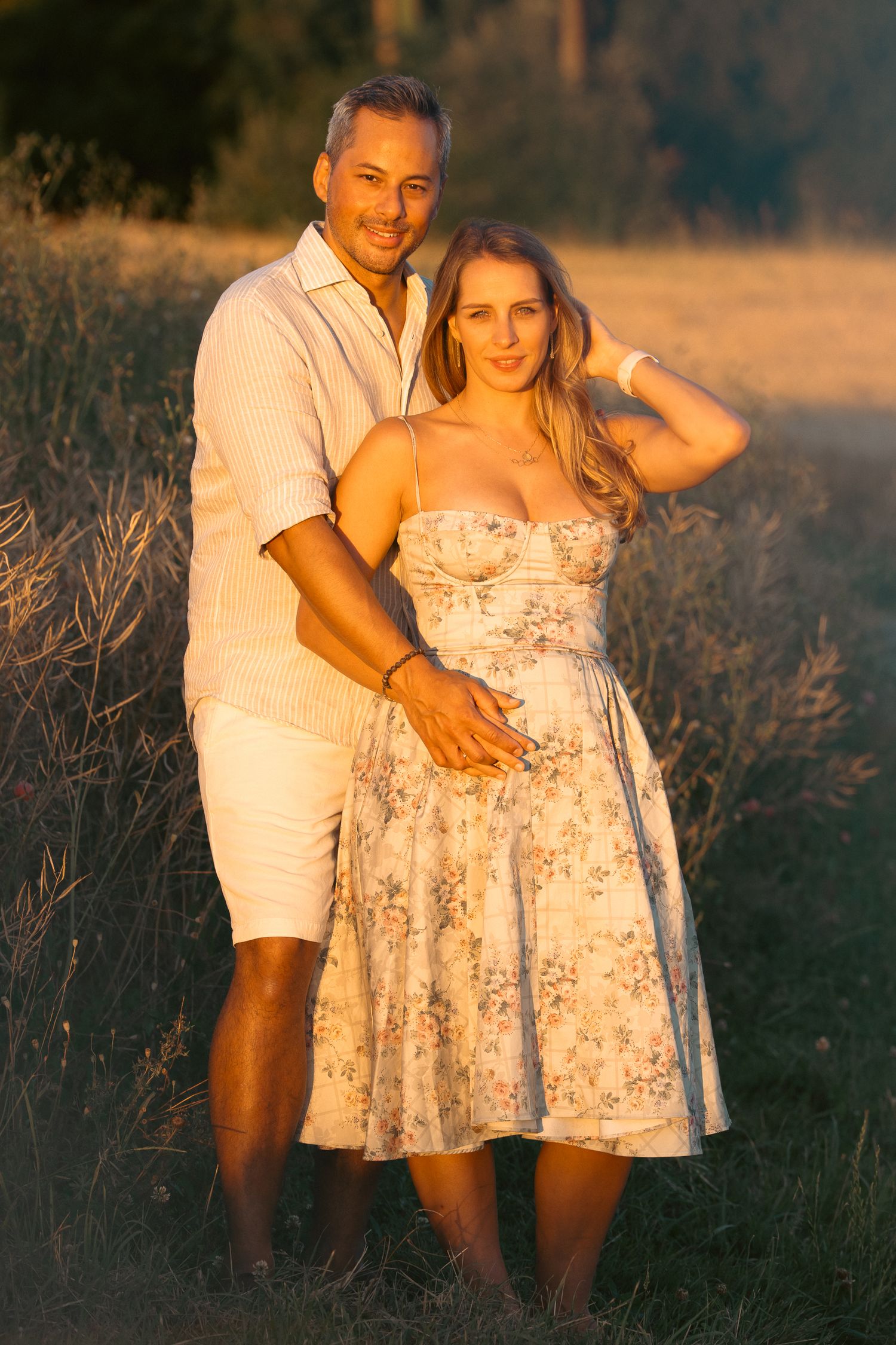 Couple posing together at golden hour in a field, woman in floral sundress, man in white casual outfit.