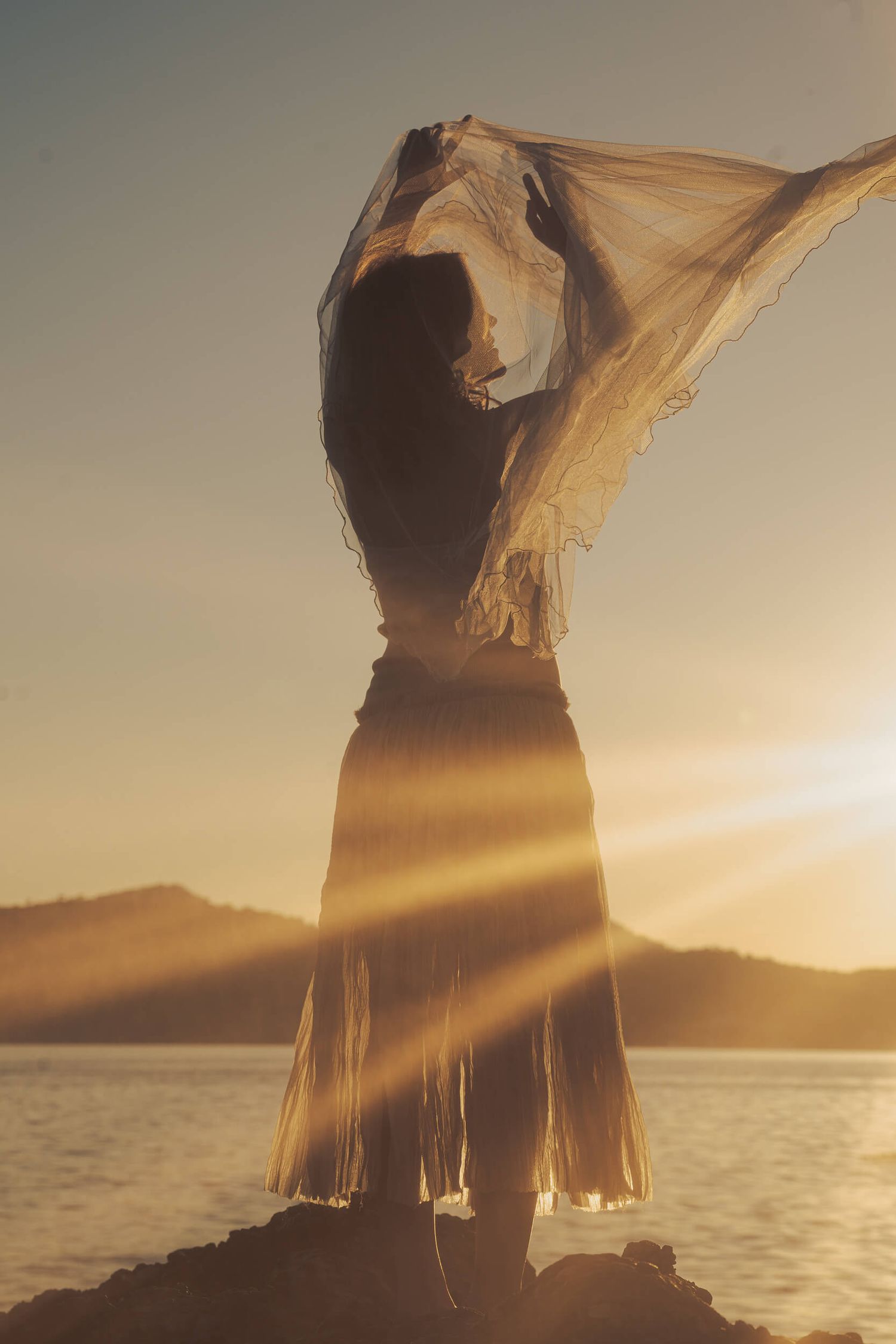 A woman in a flowing skirt holds a sheer fabric aloft against a golden sunset over calm water.
