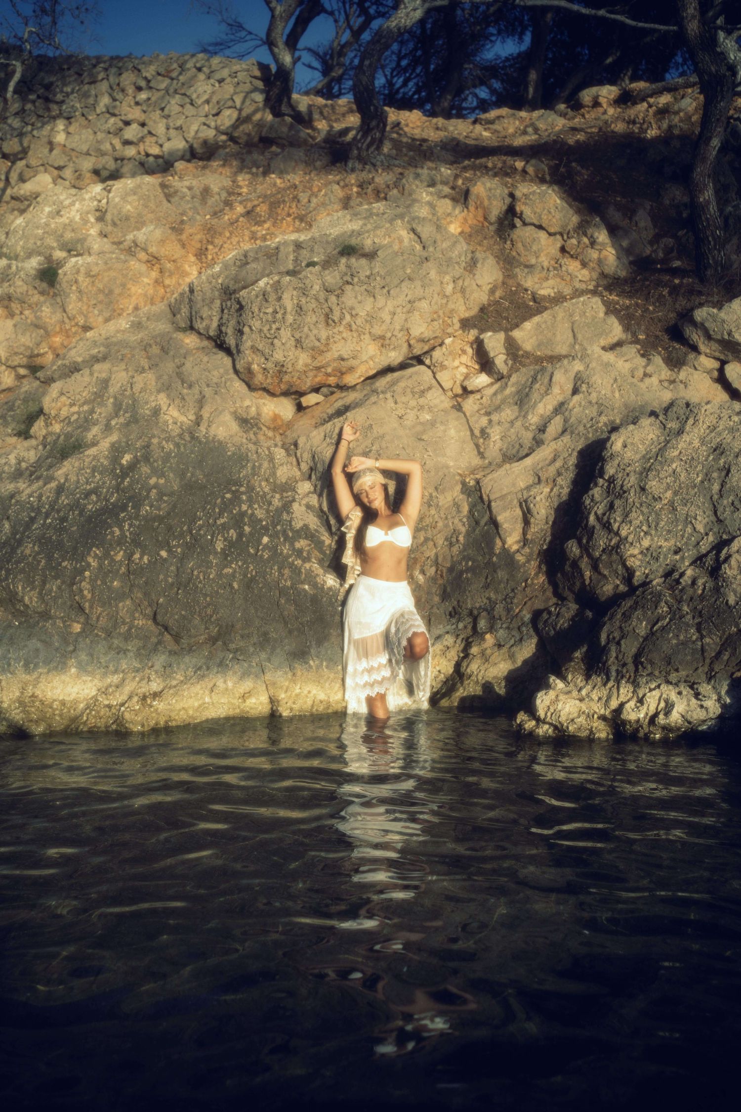 Woman in white bikini top and skirt standing in water against rocky cliff backdrop.
