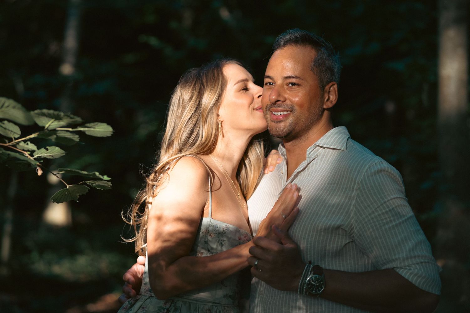 A woman kisses a smiling man on the cheek outdoors, surrounded by lush green trees.