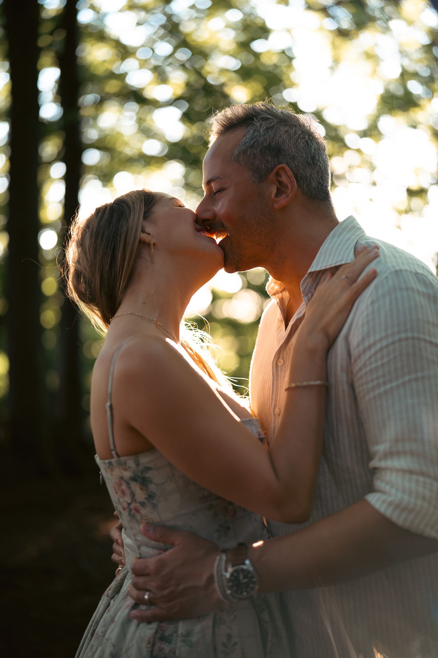 Romantic couple sharing a kiss outdoors in golden sunlight with tall trees in the background.