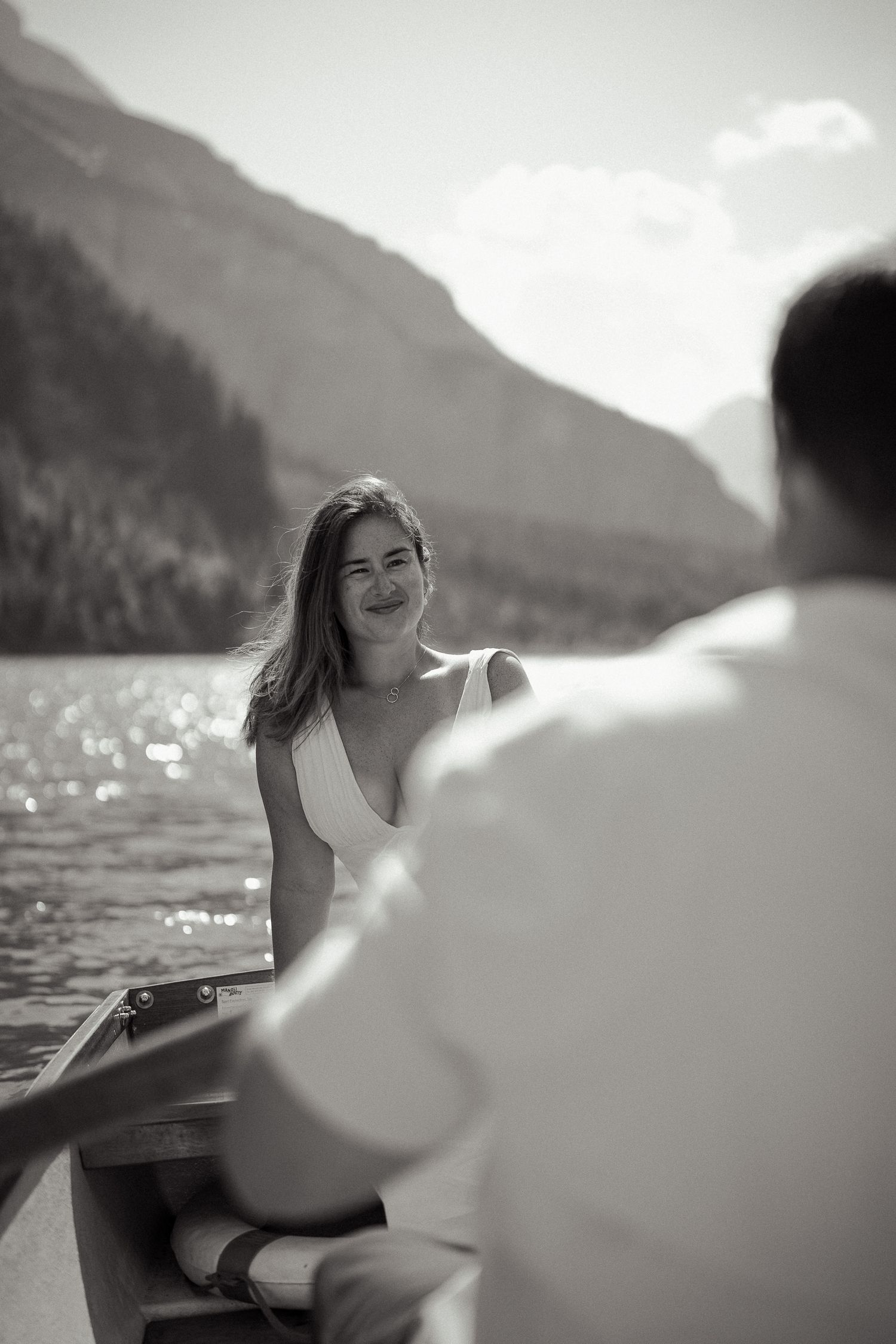 Black and white photo of a smiling bride in a white dress sitting in a boat on a mountain lake.