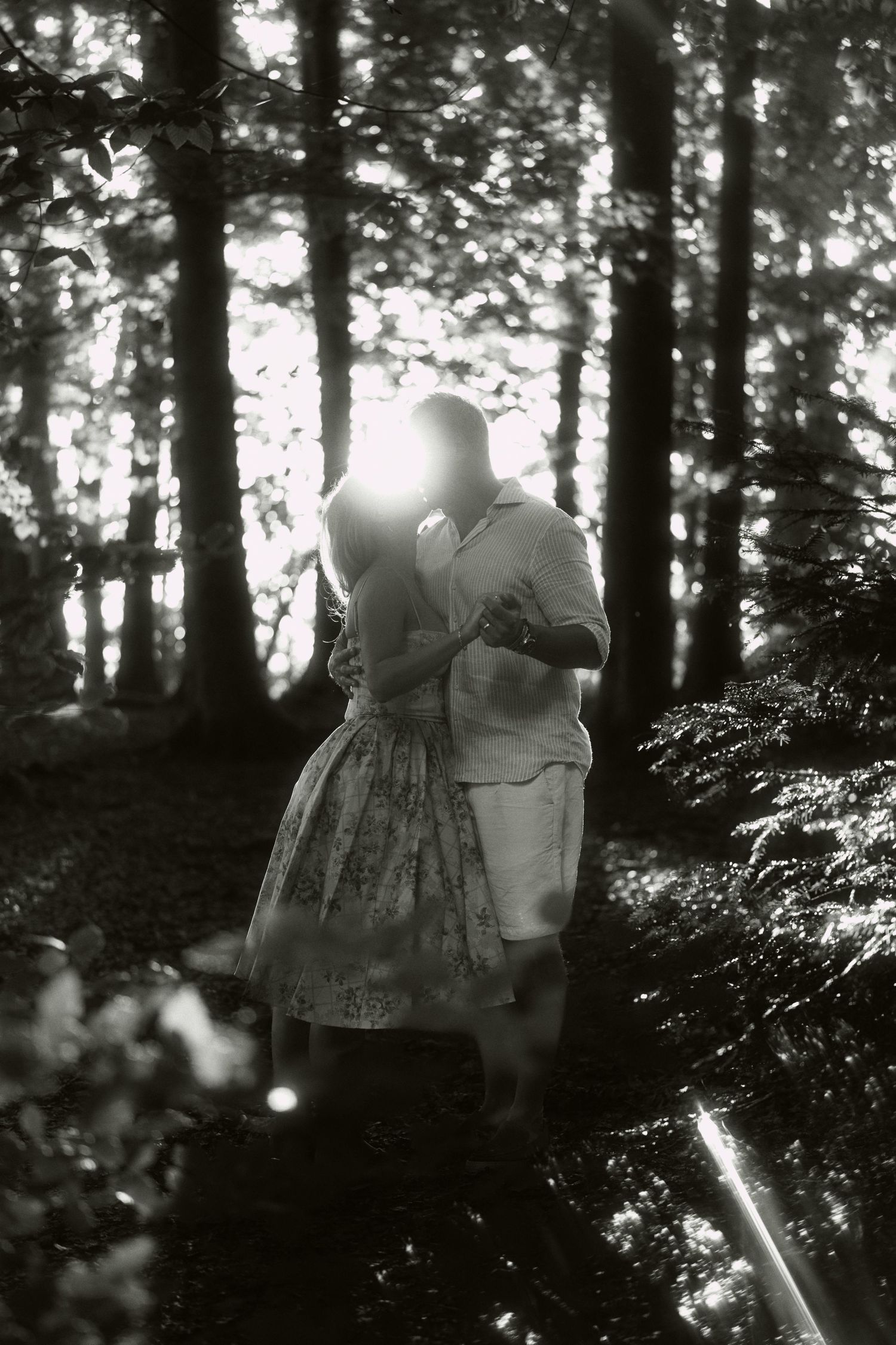Black and white photo of a couple embracing romantically in a sunlit forest, surrounded by tall trees and dappled light.