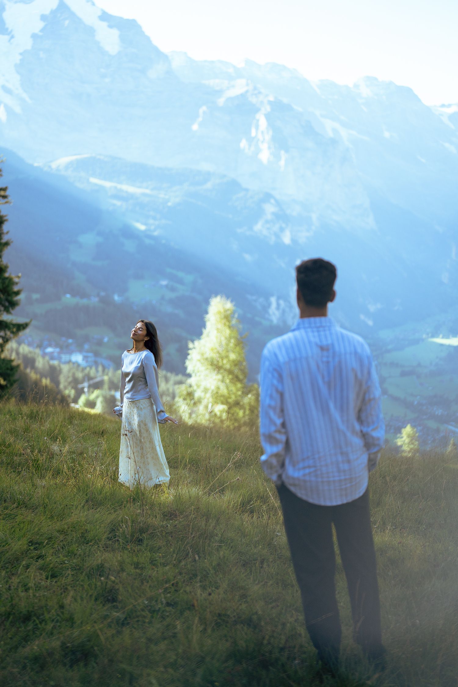 Couple standing on a grassy alpine meadow with majestic snow-capped mountains in the background on a sunny day.