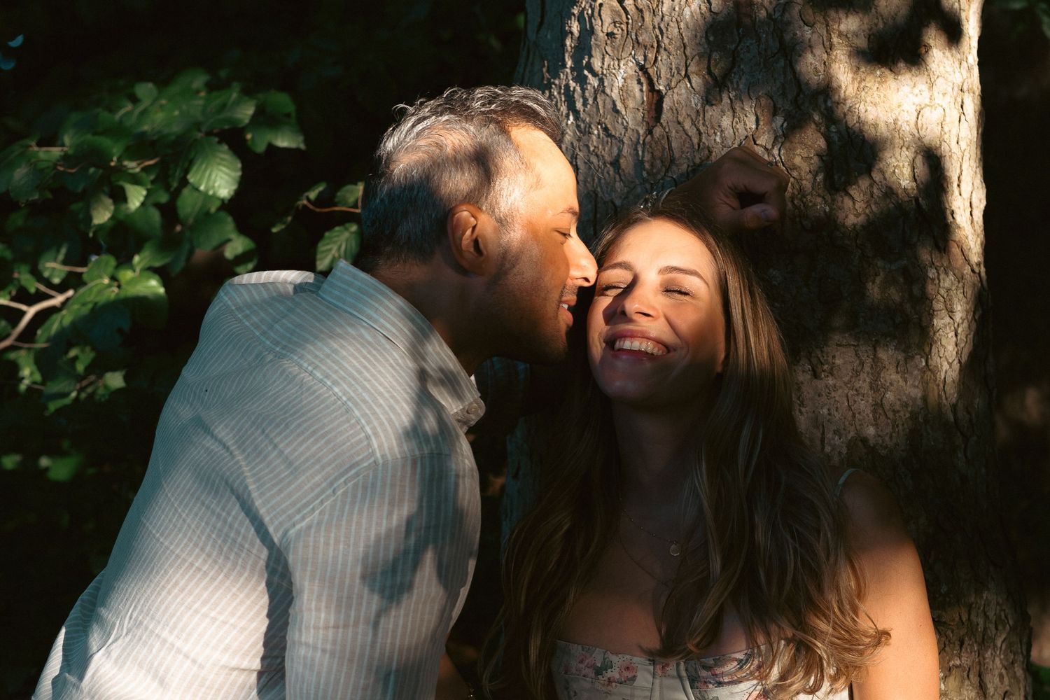 Man kissing woman on the cheek as she laughs joyfully against a tree in a forest setting.
