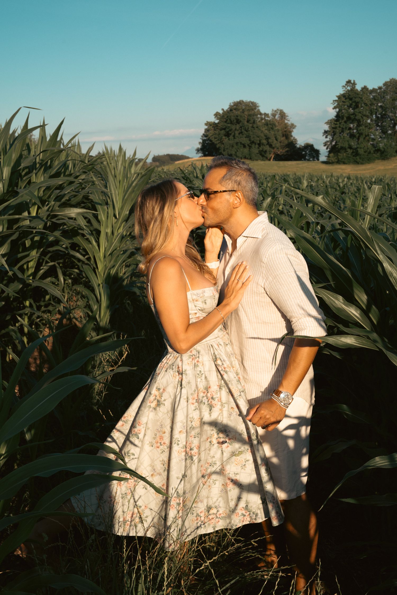 Couple sharing a romantic kiss in a sunlit cornfield, woman in floral dress, man in white shirt wearing a watch.