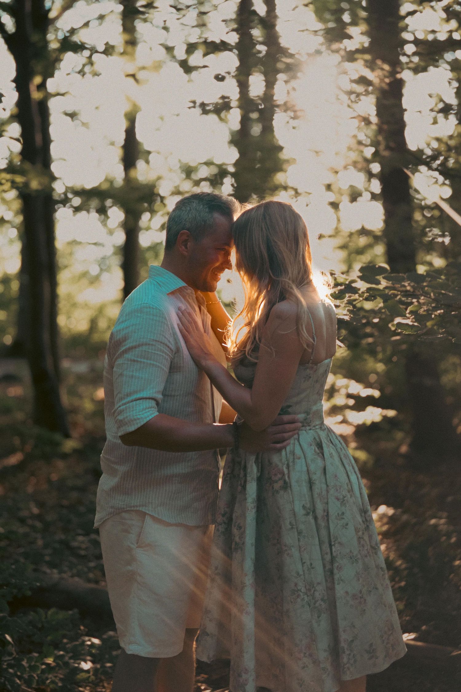 Couple embracing in a sunlit forest, sharing a romantic moment among trees with golden hour light.