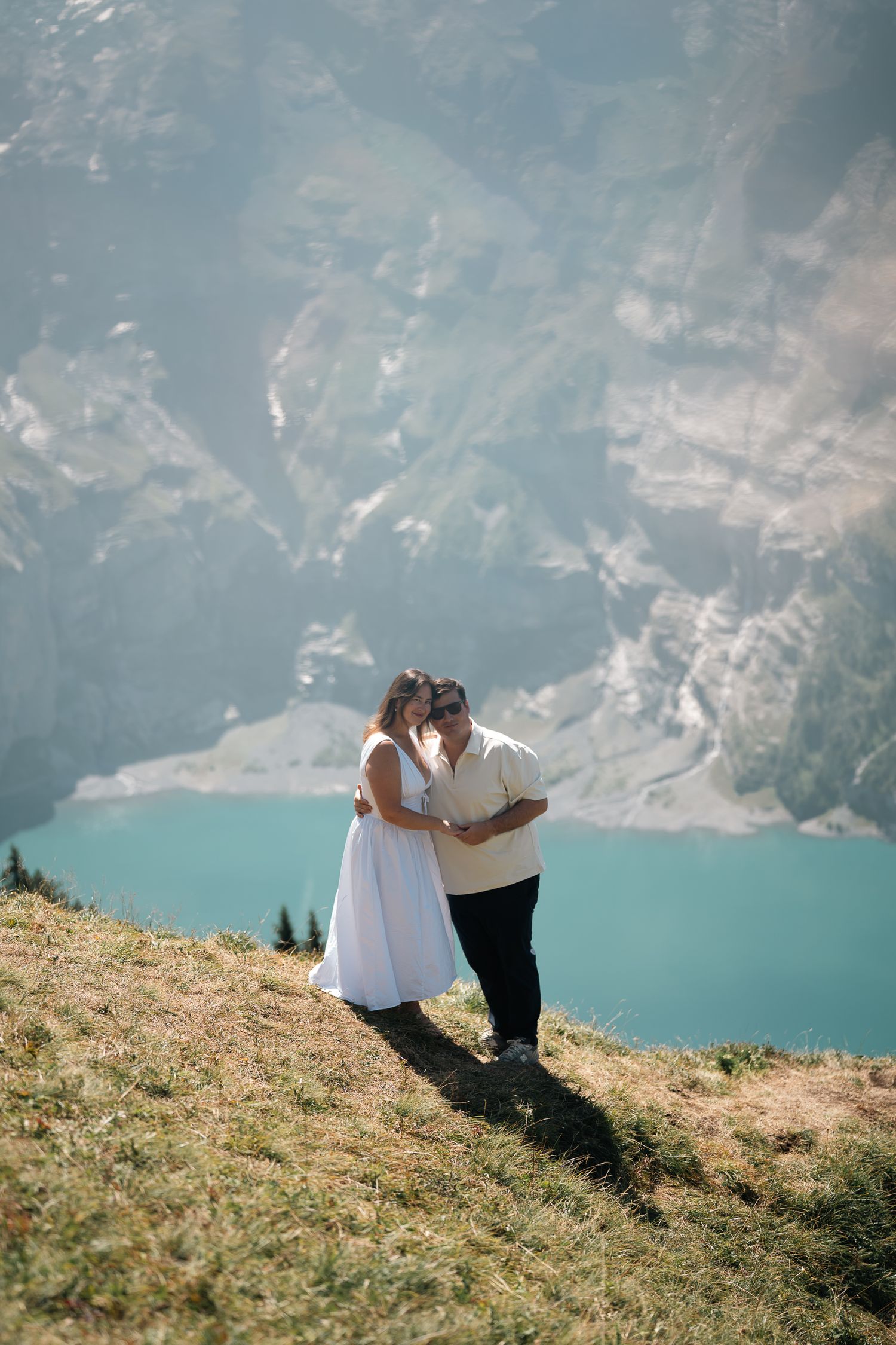 Couple embracing on a mountain hillside with a turquoise alpine lake and rocky peaks in the background.