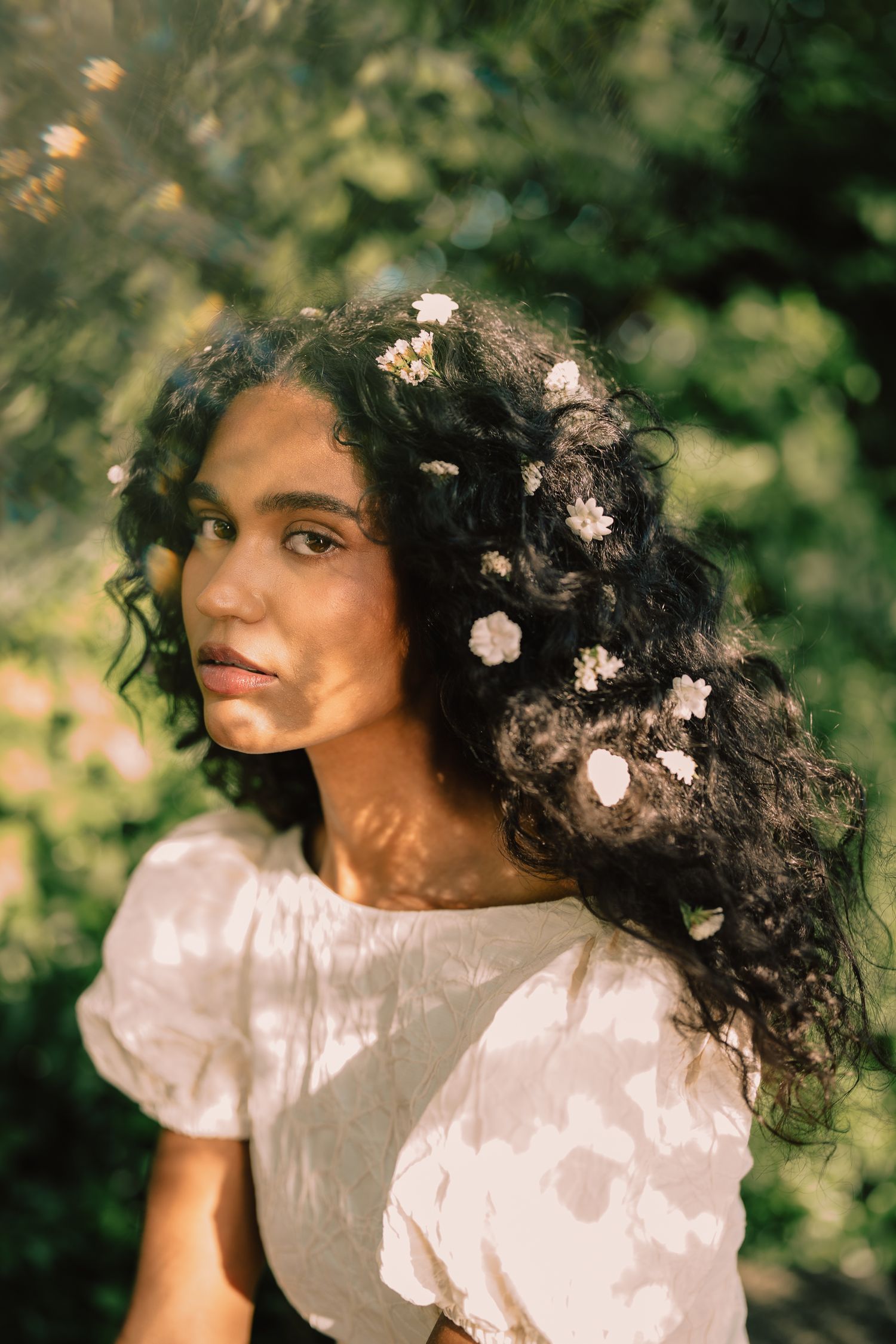 Young woman with curly dark hair adorned with small white flowers, wearing a white blouse outdoors.