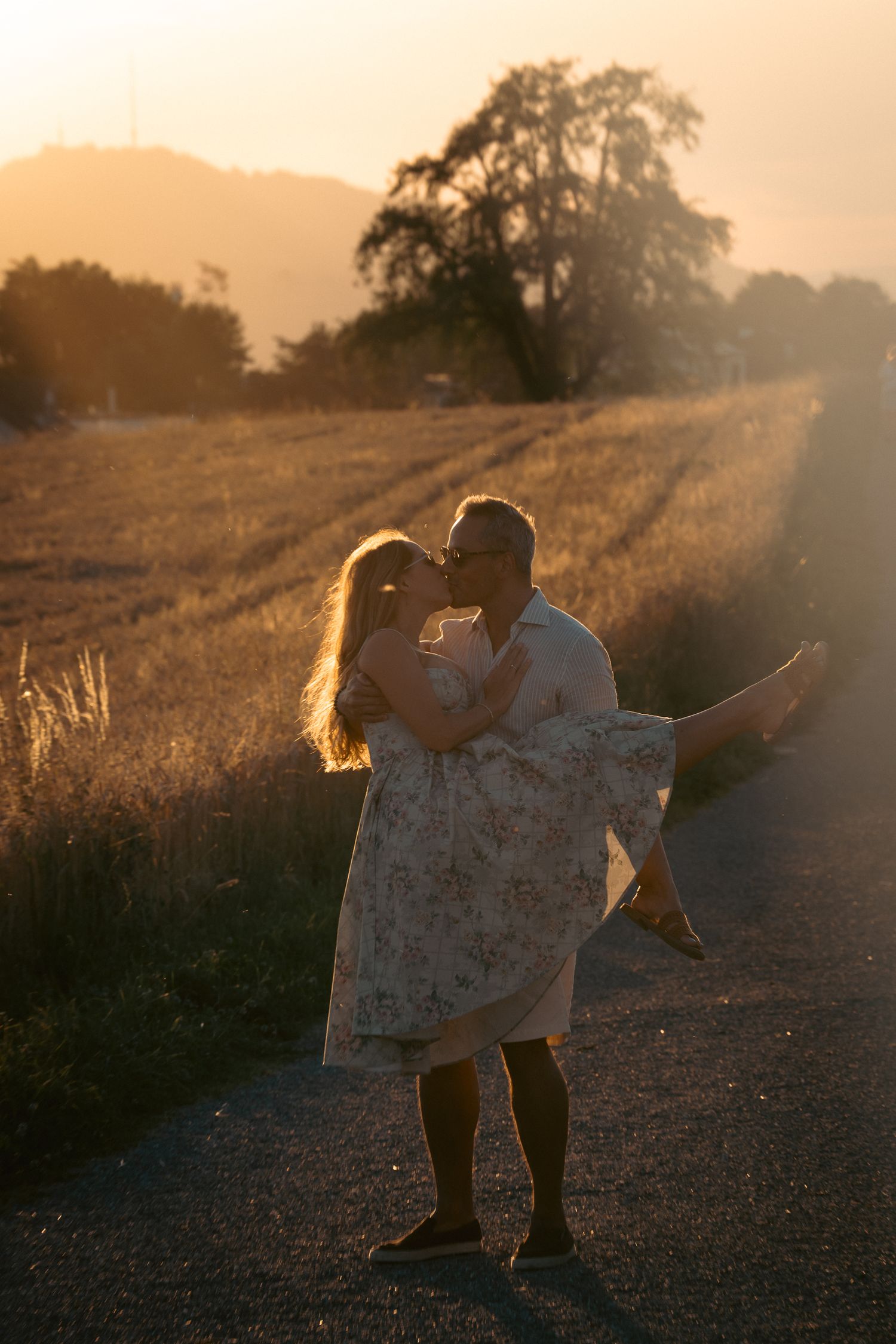 Romantic couple kissing at sunset on country road, man carrying woman in floral dress with golden field backdrop.