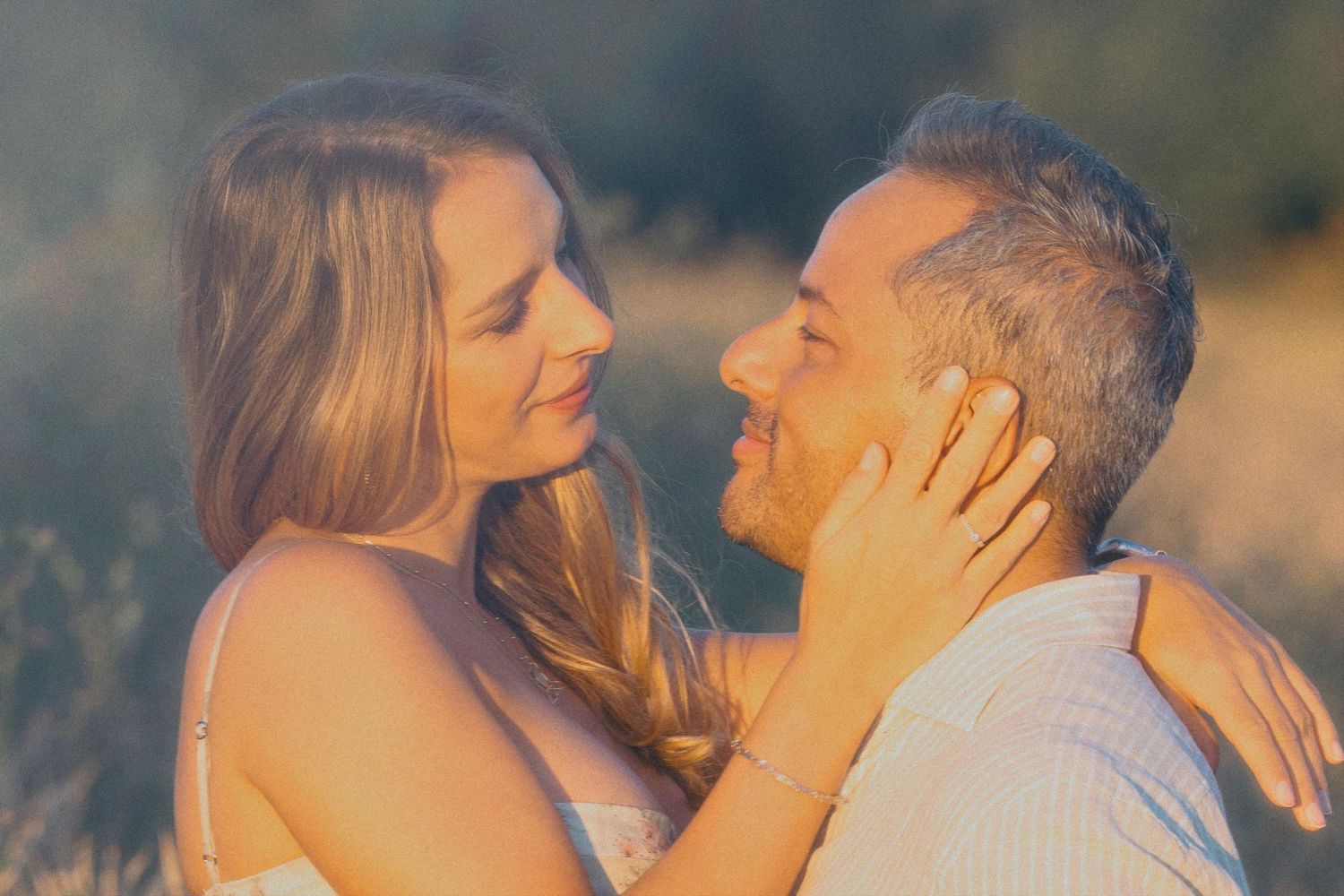 A couple shares a tender romantic moment outdoors, the woman gently cupping the man's face in warm golden light.