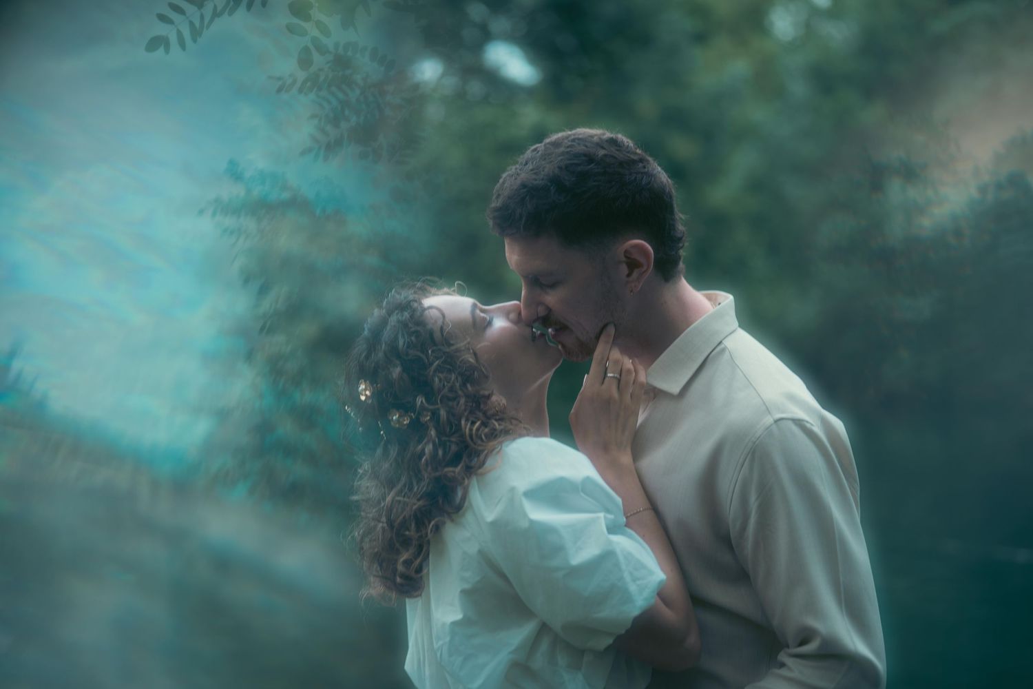 A couple leans in for a kiss surrounded by lush green foliage and soft light.