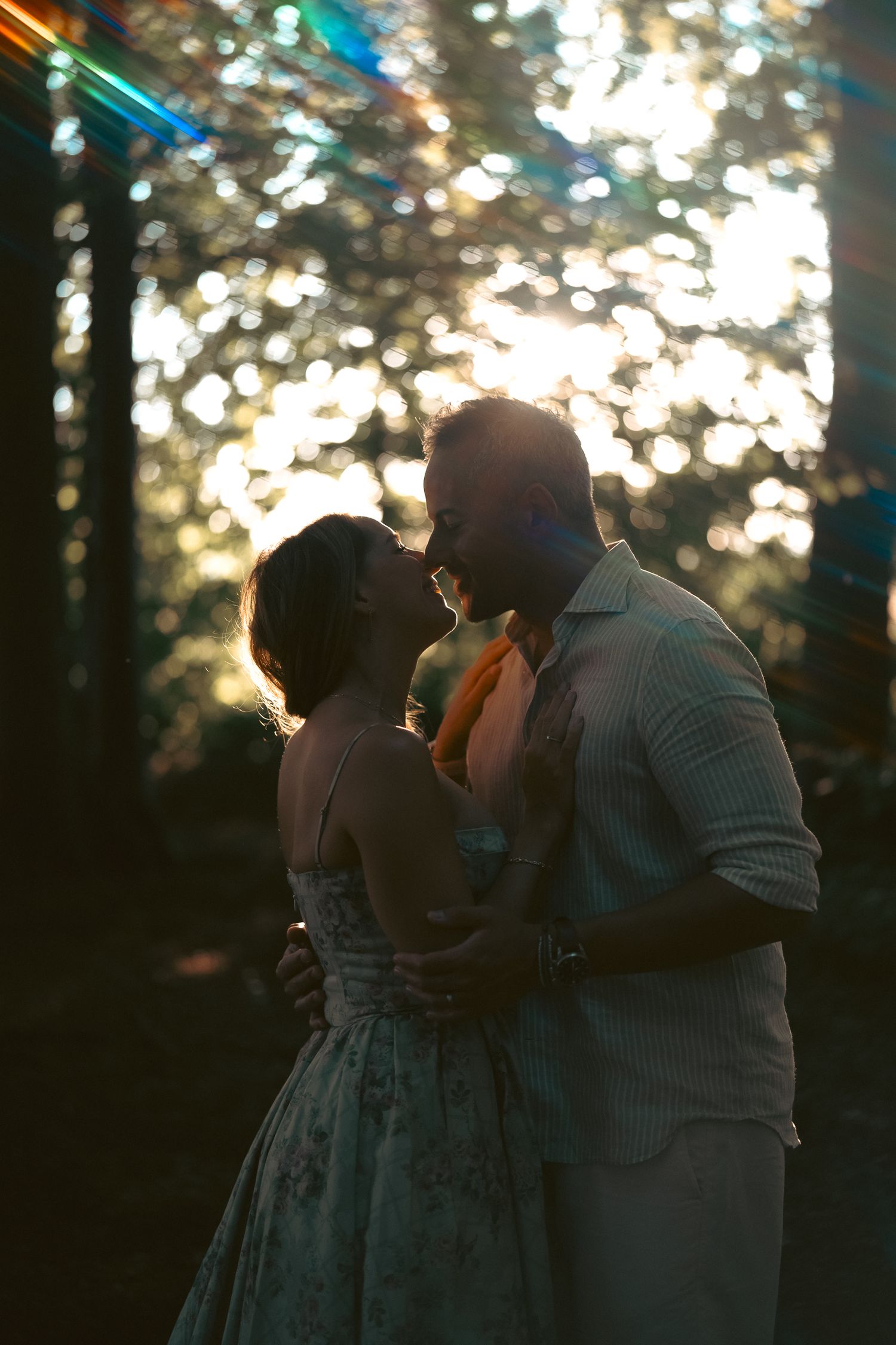 A couple embraces and kisses in silhouette against a stunning bokeh forest backdrop with golden glowing light.