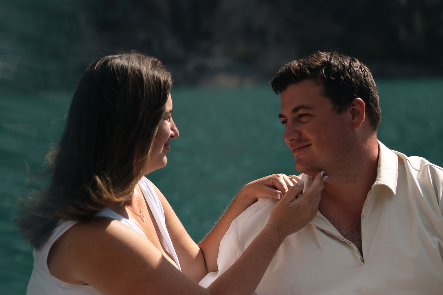 A couple shares a tender moment outdoors near turquoise water, woman touching man's shirt collar.