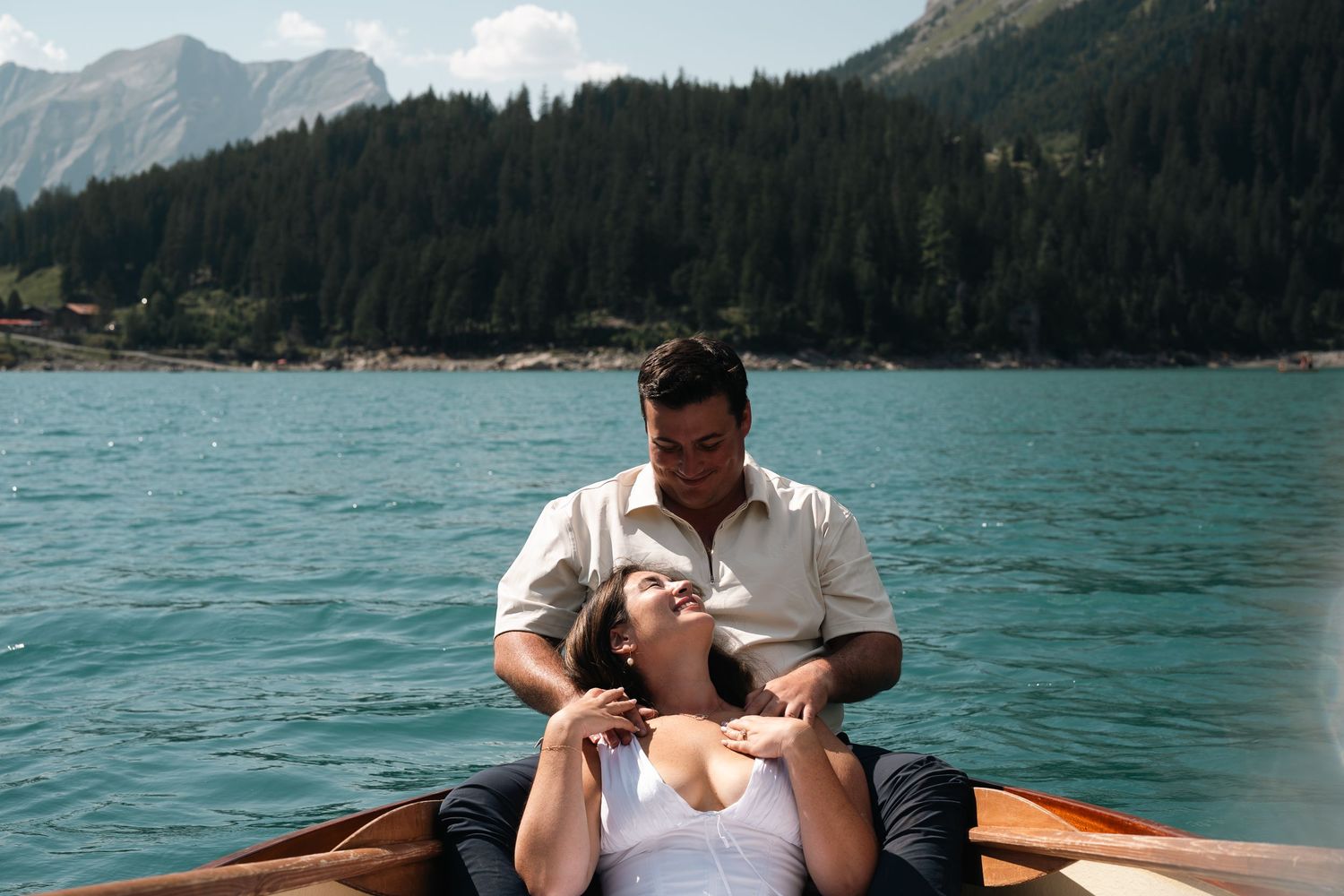 Couple relaxing in a wooden rowboat on a turquoise alpine lake surrounded by forested mountains.