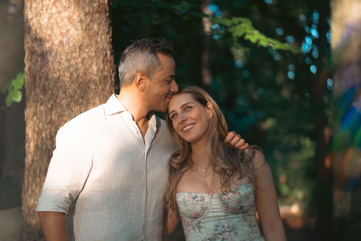 Couple sharing a romantic moment among trees in warm sunlight, man kissing woman's head as she smiles up at him.