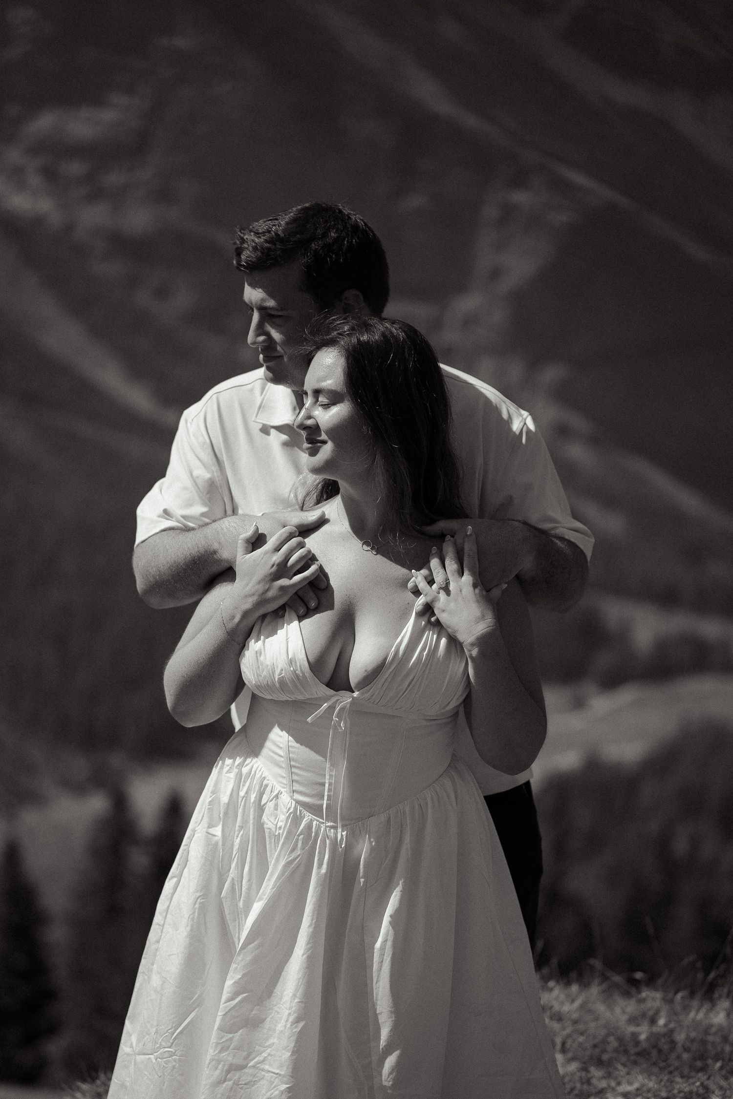 Black and white photo of a couple embracing in a romantic pose with mountain scenery in the background.