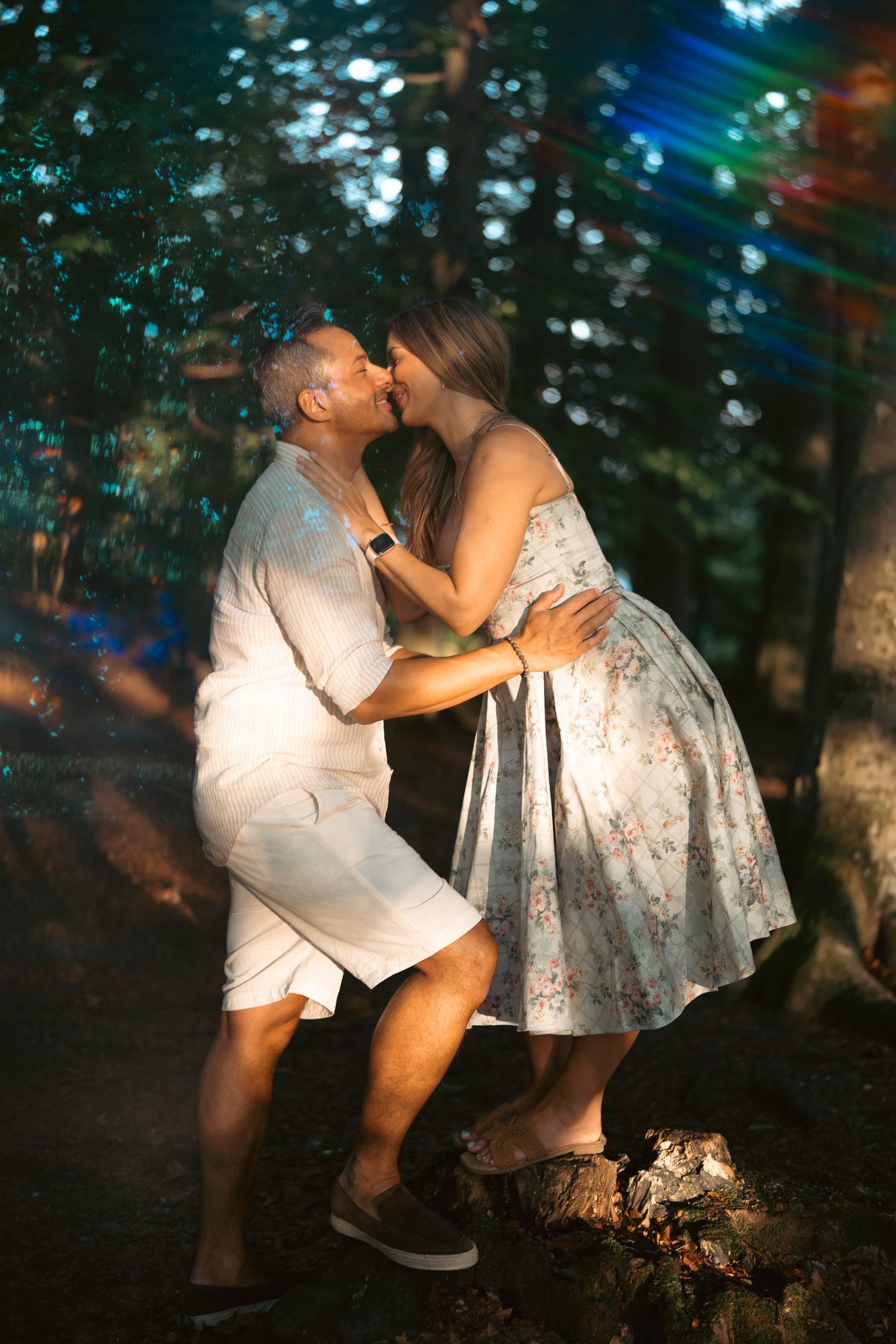 Couple sharing a romantic kiss in a sunlit forest, man in white outfit and woman in floral dress.