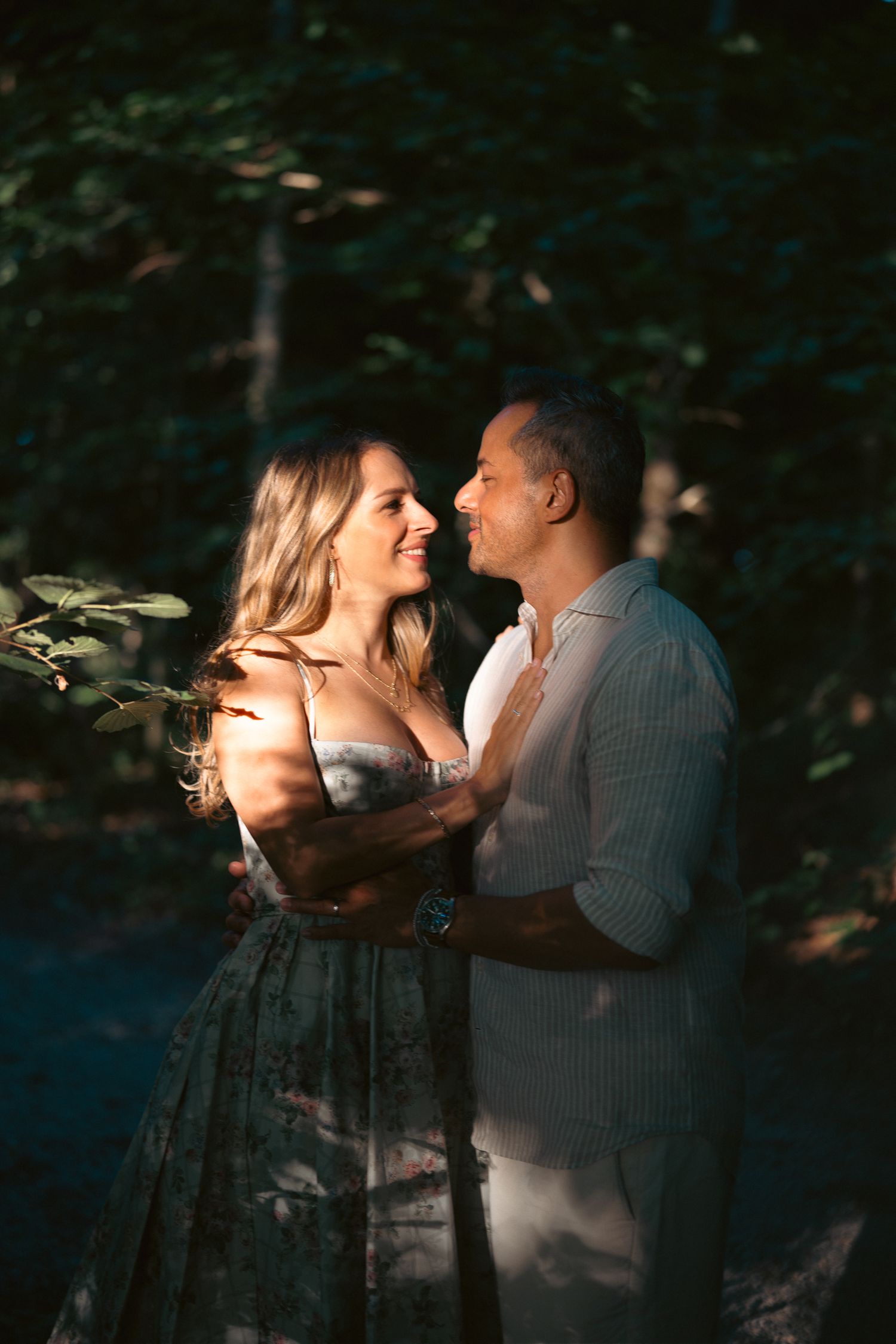 Couple embracing and smiling at each other in a sunlit forest, woman in floral dress, man in light shirt.
