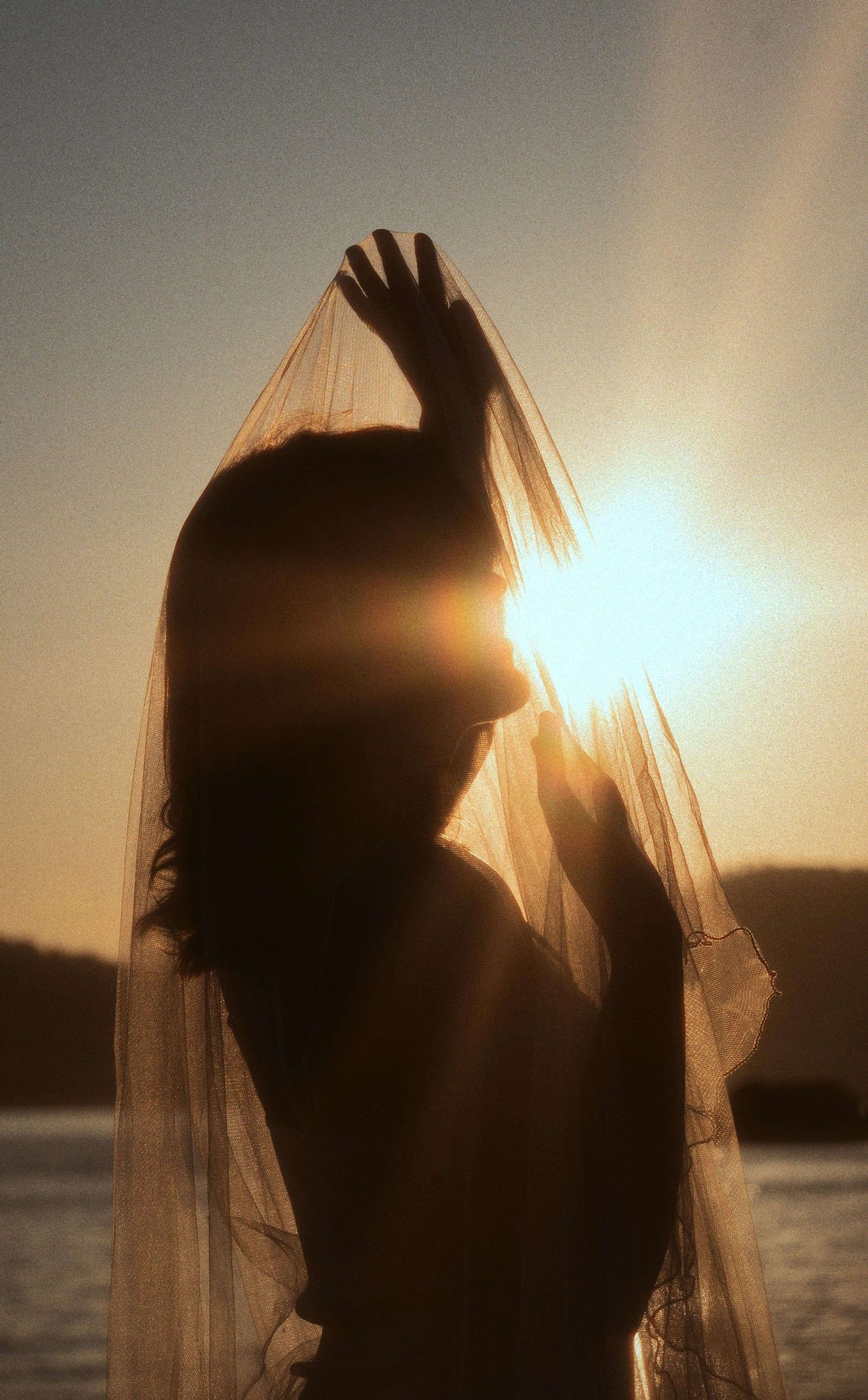 Silhouette of a woman holding a sheer veil against a golden sunset over water, backlit by glowing sunlight.