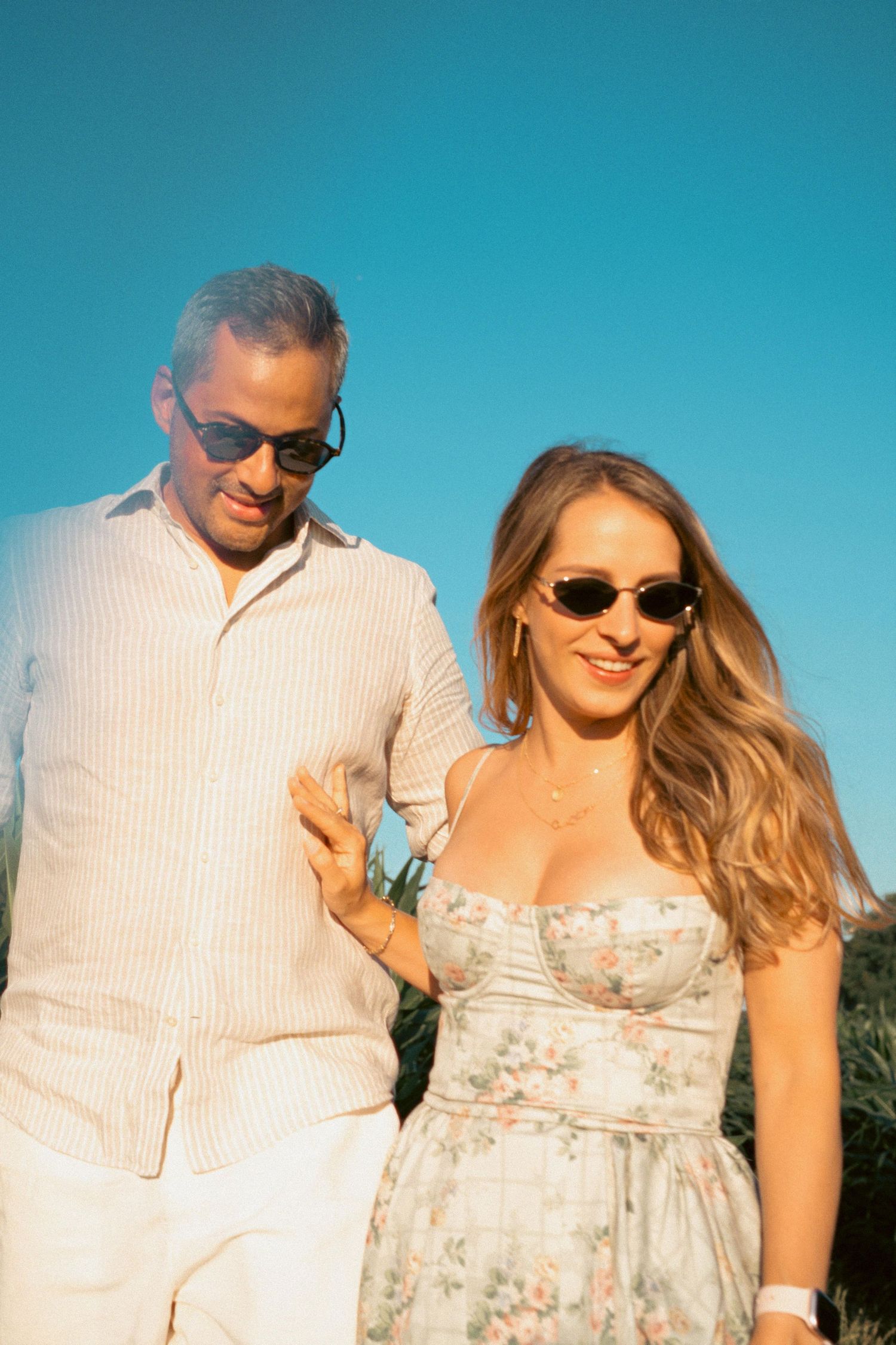 Couple in sunglasses walking outdoors under a clear blue sky, woman in floral dress, man in white linen shirt.